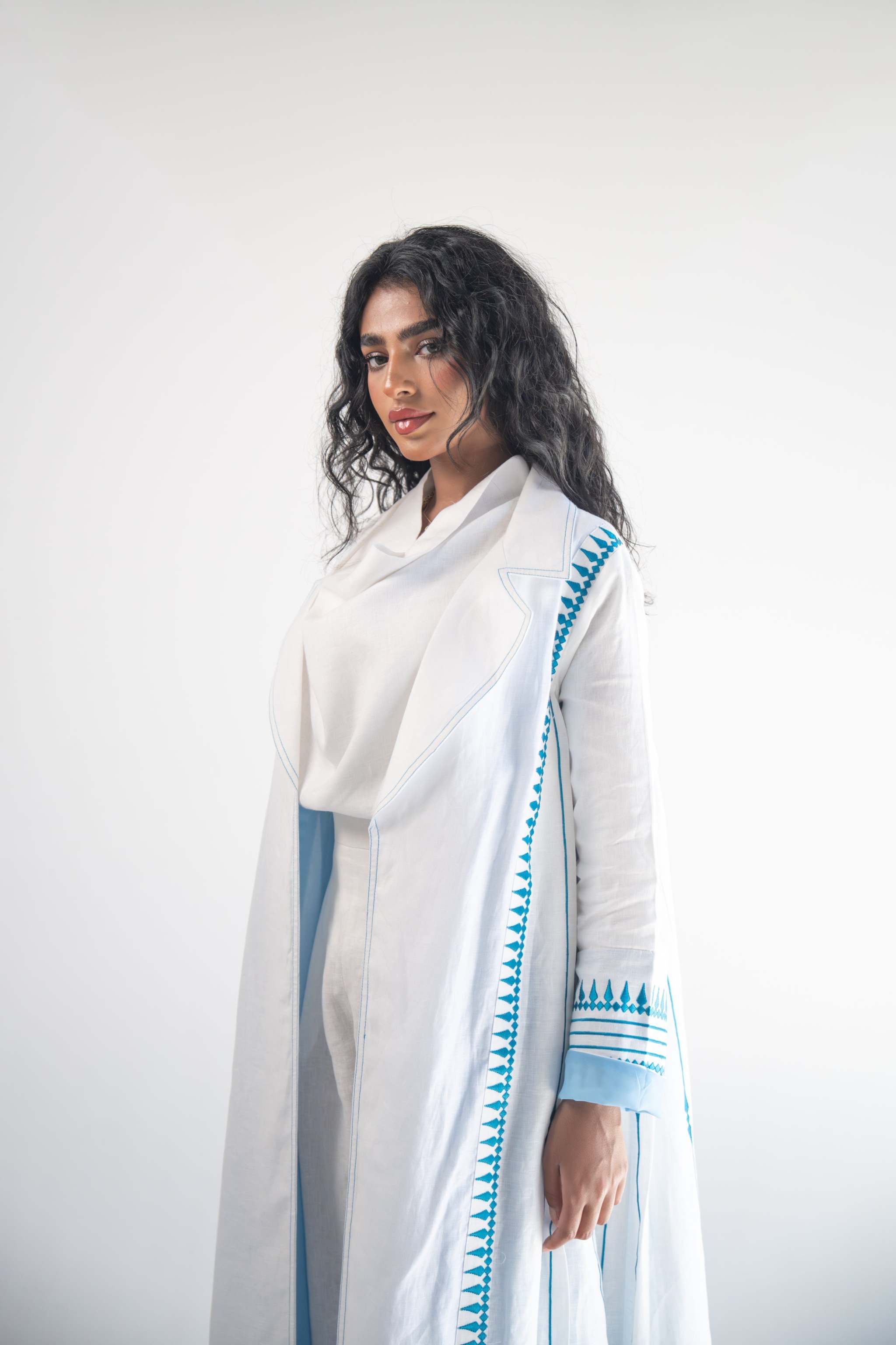 Woman wearing white linen blazer abaya with blue triangular embroidery and chiffon inner layer.
