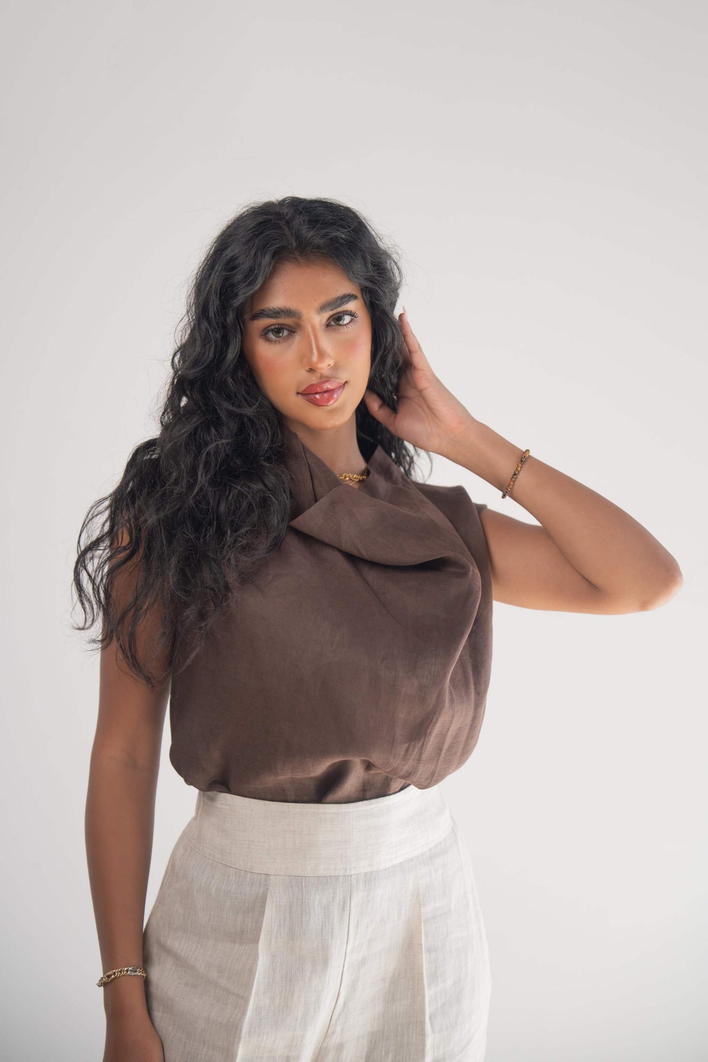 Woman wearing a sleeveless cocoa linen blouse with a draped neckline and white linen pants against a plain background