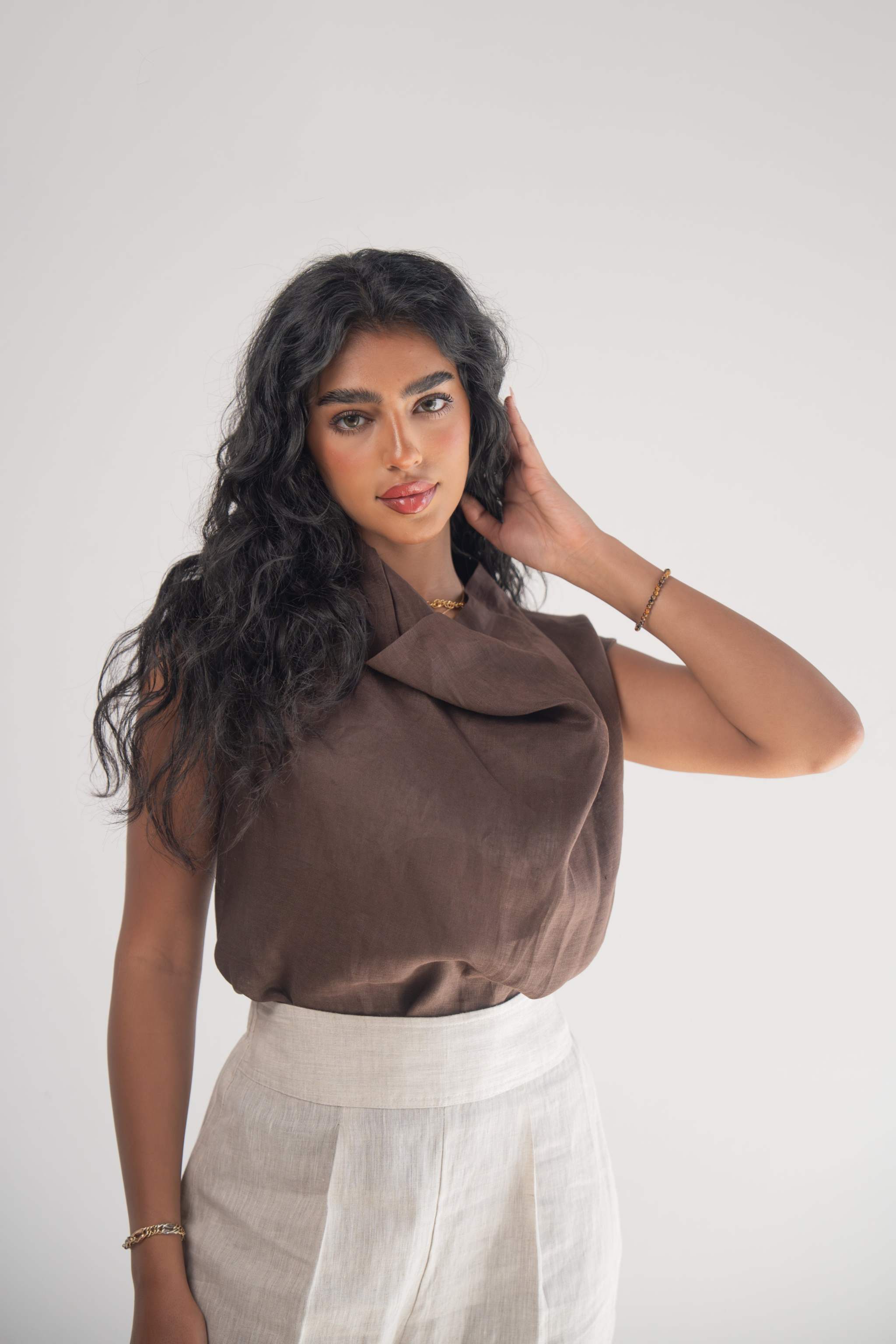 Woman wearing a sleeveless cocoa linen blouse with a draped neckline and white linen pants against a plain background