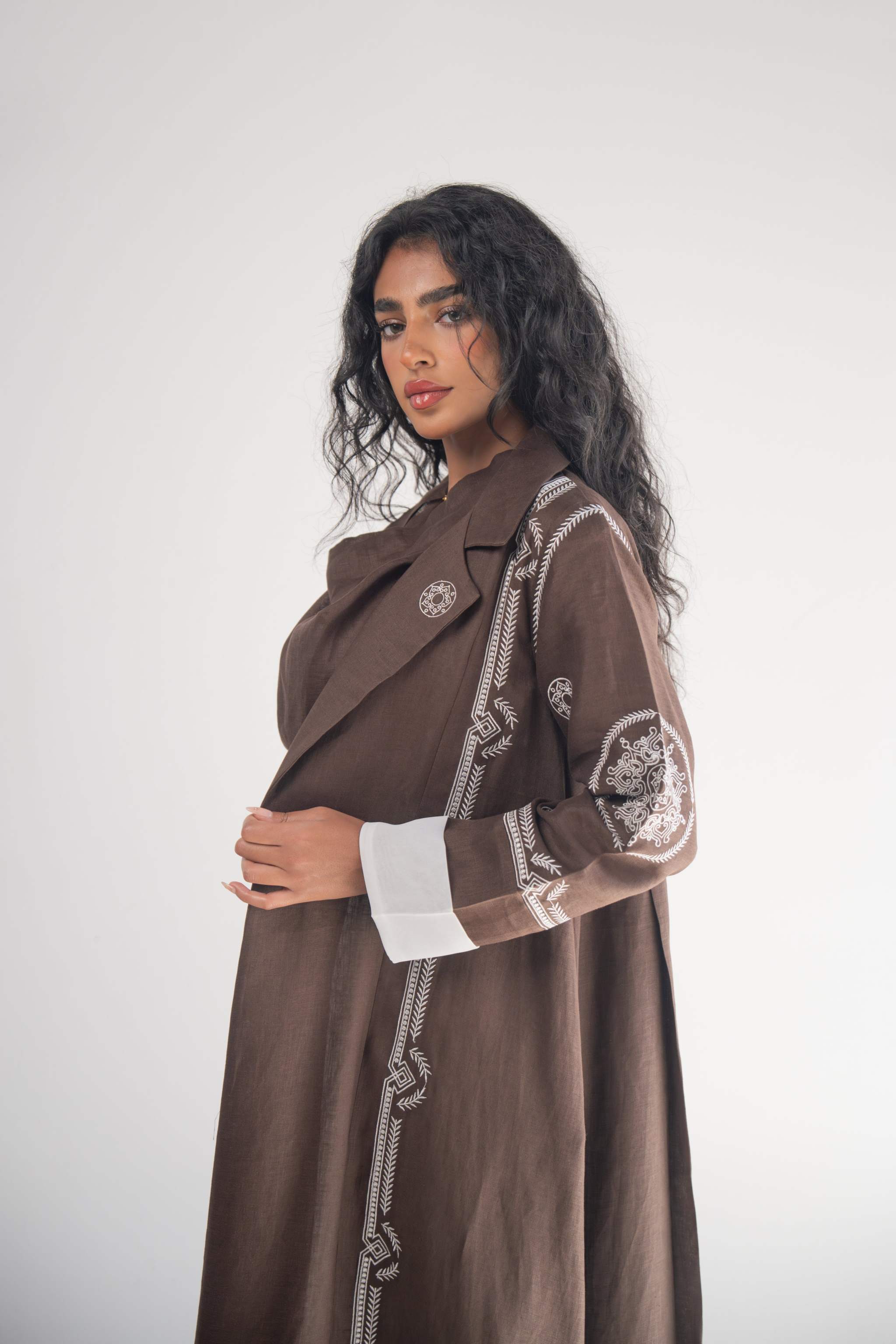 Woman wearing a cocoa brown linen blazer abaya with white geometric and circular embroidery on sleeves and sides, featuring a classic blazer lapel and white chiffon lining.