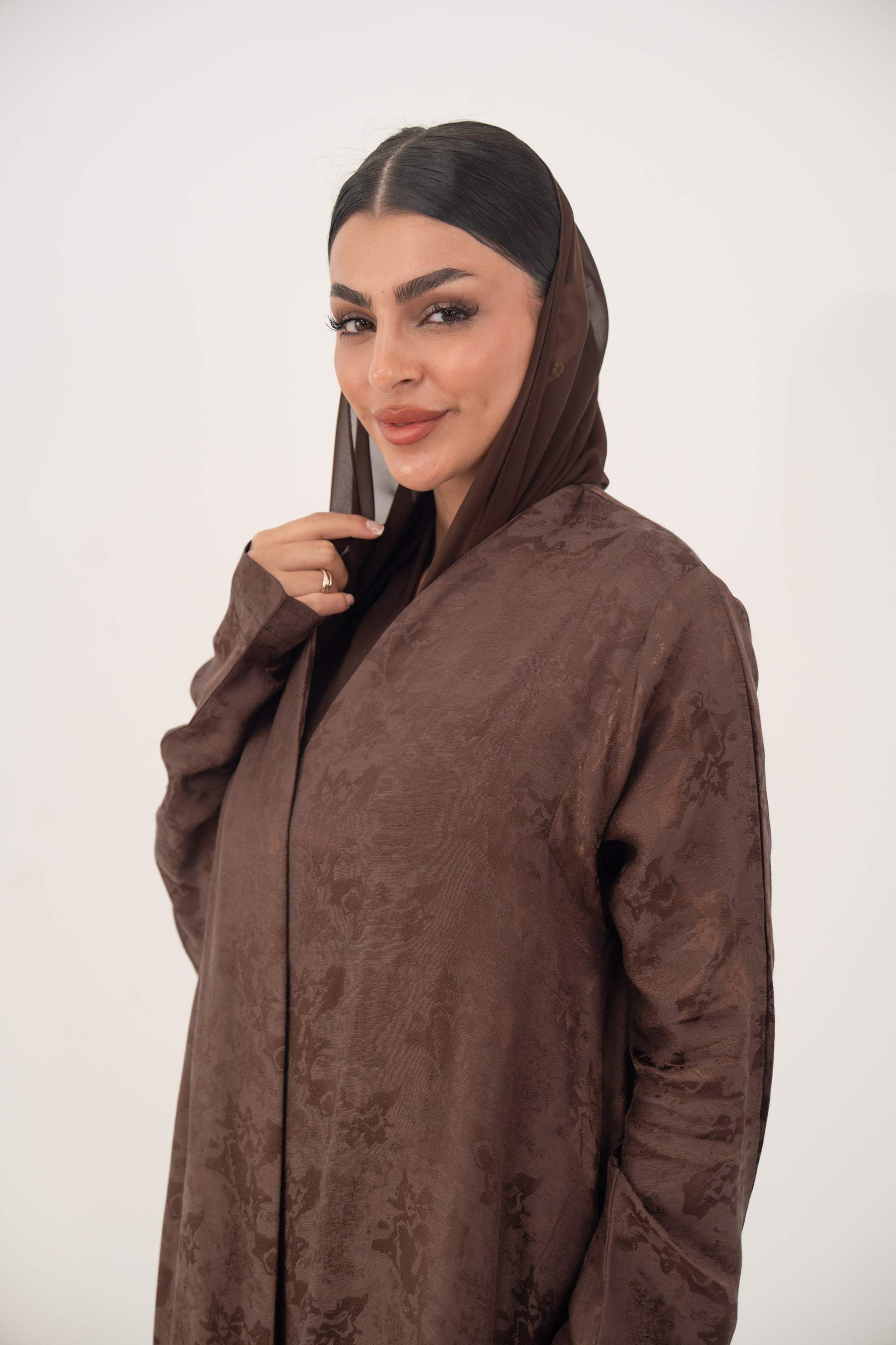 Woman wearing a warm cocoa brown jacquard silk abaya with subtle floral pattern and long sleeves, featuring a relaxed A-line cut and clean edges.