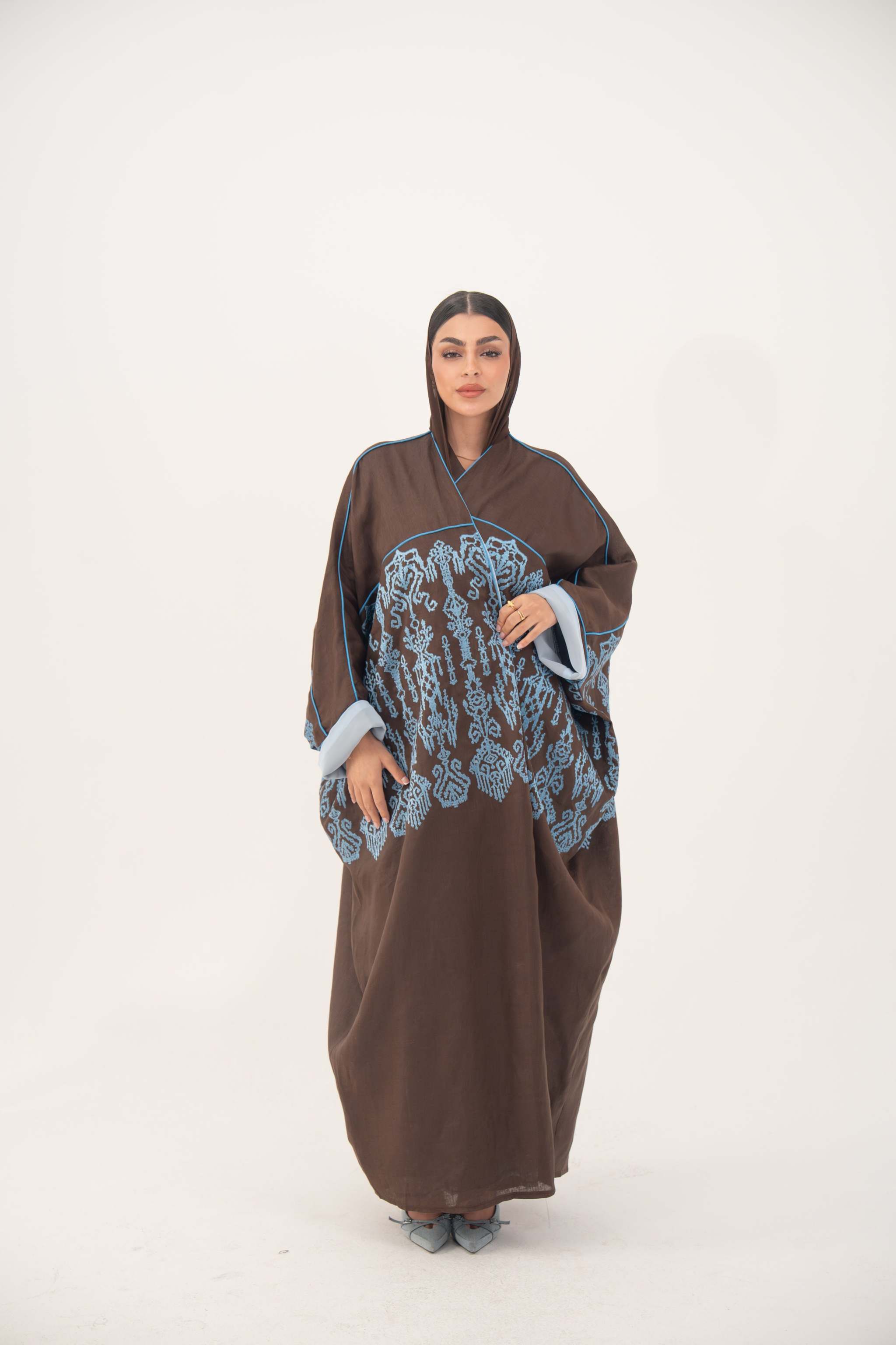 Woman wearing a loose mocha brown bisht abaya with blue shak embroidery on back and sleeves, light blue piping, and rolled cuffs.