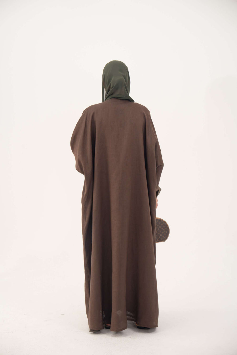 Back view of a brown bisht abaya with loose elegant cut, worn with a dark green hijab and holding a small handbag