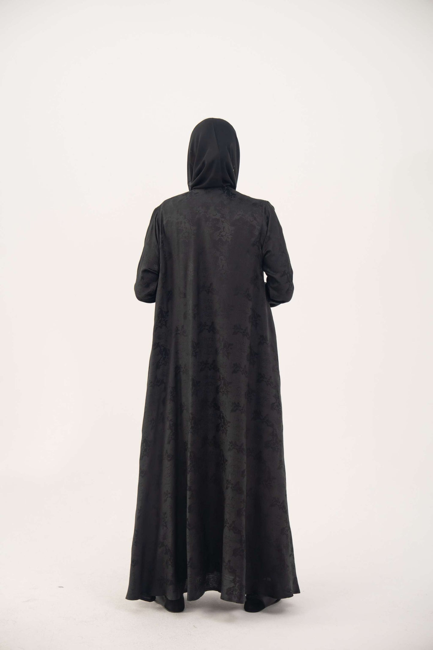 Back view of a black jacquard silk abaya with tone-on-tone floral weave, featuring a fluid A-line cut, relaxed shoulders, open front, straight sleeves, and discreet side pockets.