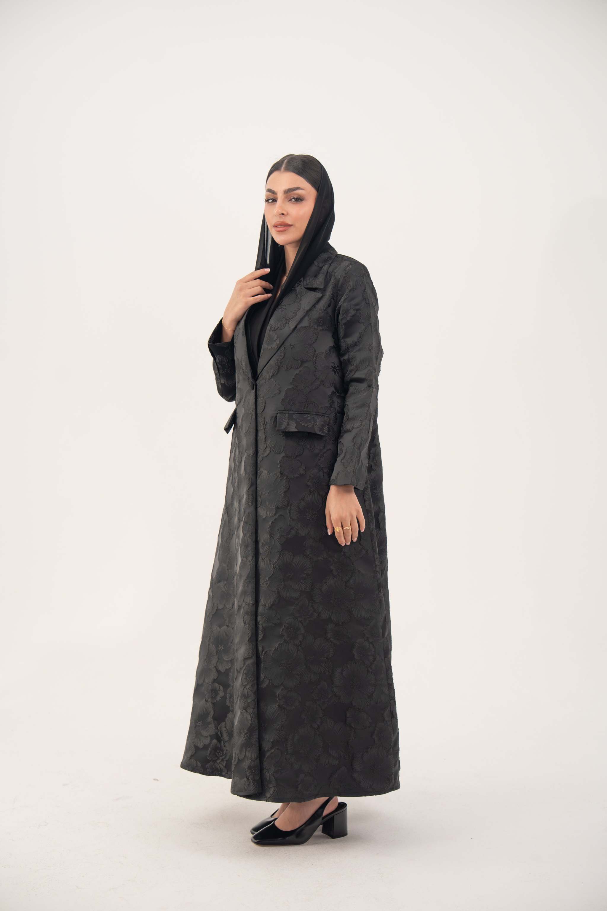 woman wearing elegant black jacquard blazer abaya with textured pattern and classic lapels standing against white background