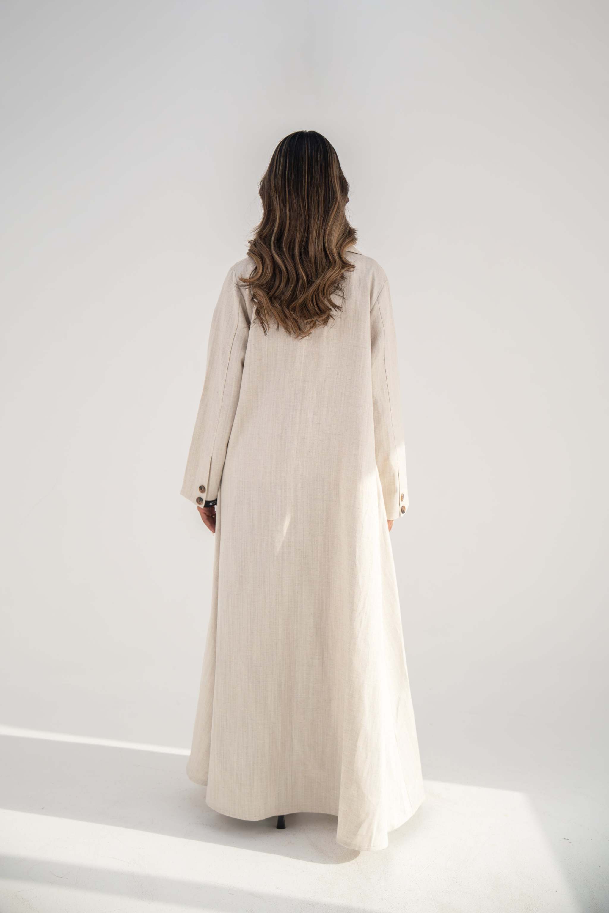 Back view of a woman wearing a warm beige blazer-style abaya made from soft twill linen blend with a flowy cut and long sleeves.