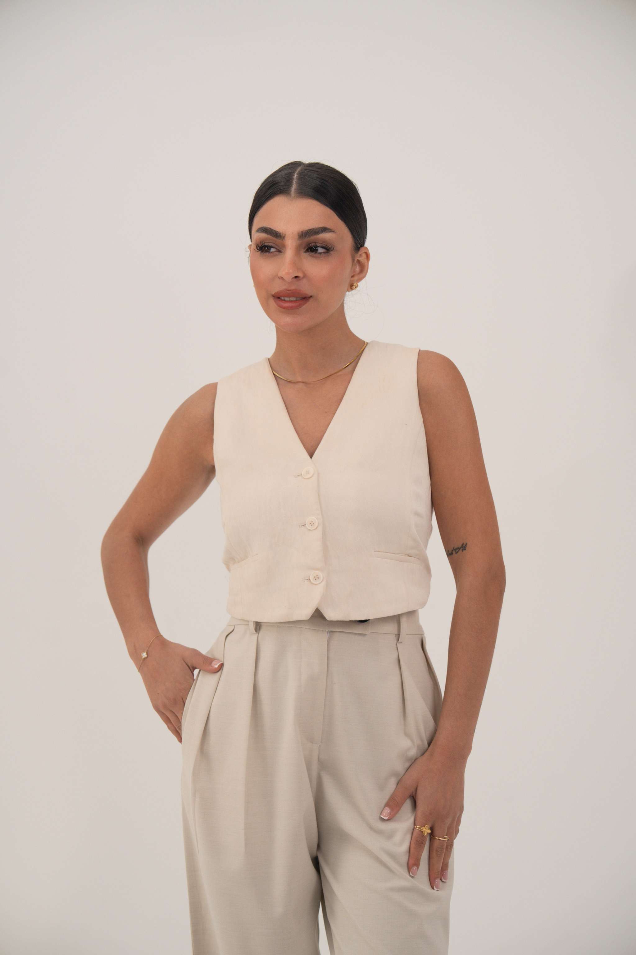 Woman wearing a soft beige sleeveless crepe vest with front buttons and light beige high-waisted pants