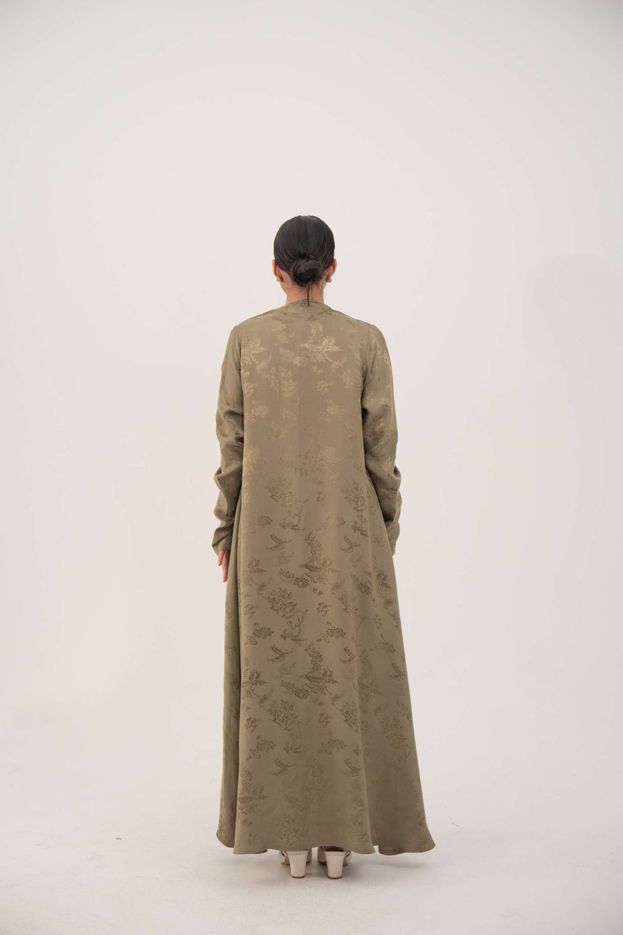 Back view of a woman wearing a soft olive green silk abaya with full-length sleeves and gentle ruching at the wrists