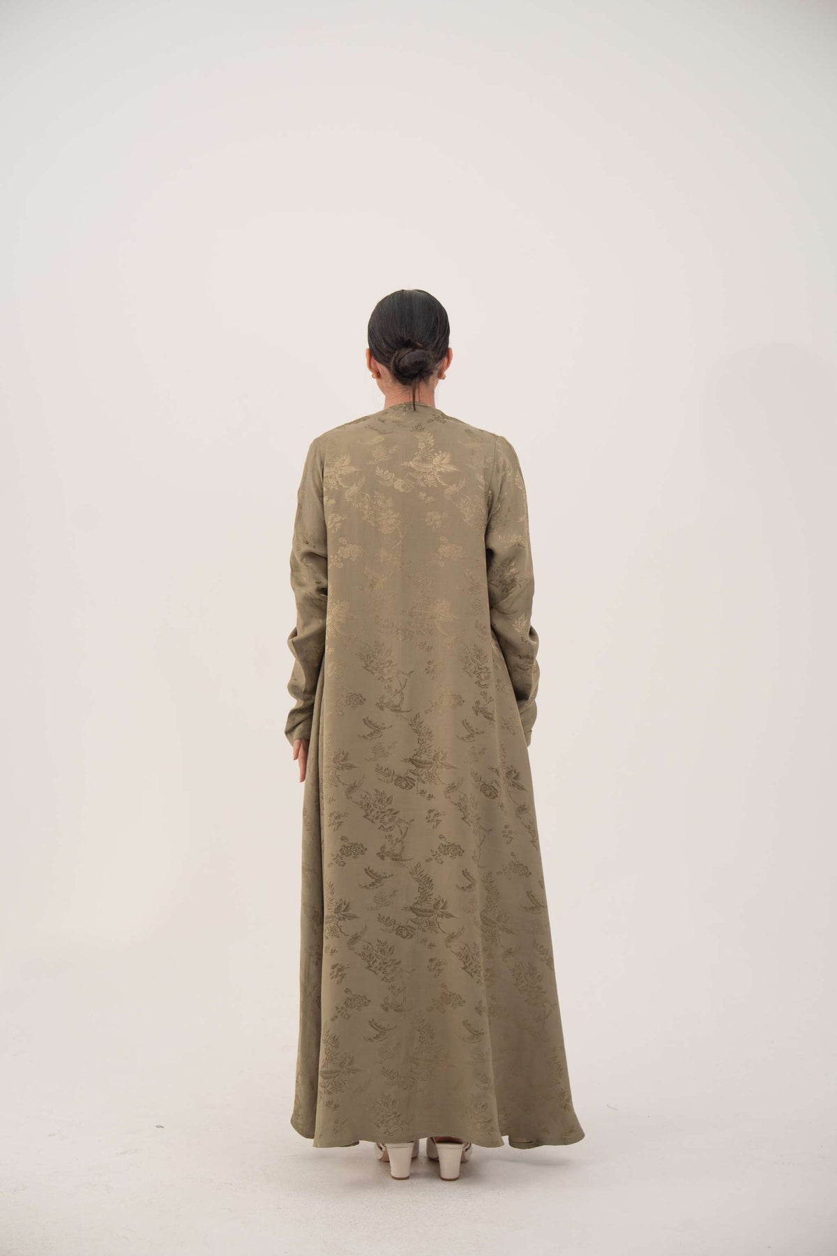 Back view of a woman wearing a soft olive green silk abaya with full-length sleeves and gentle ruching at the wrists