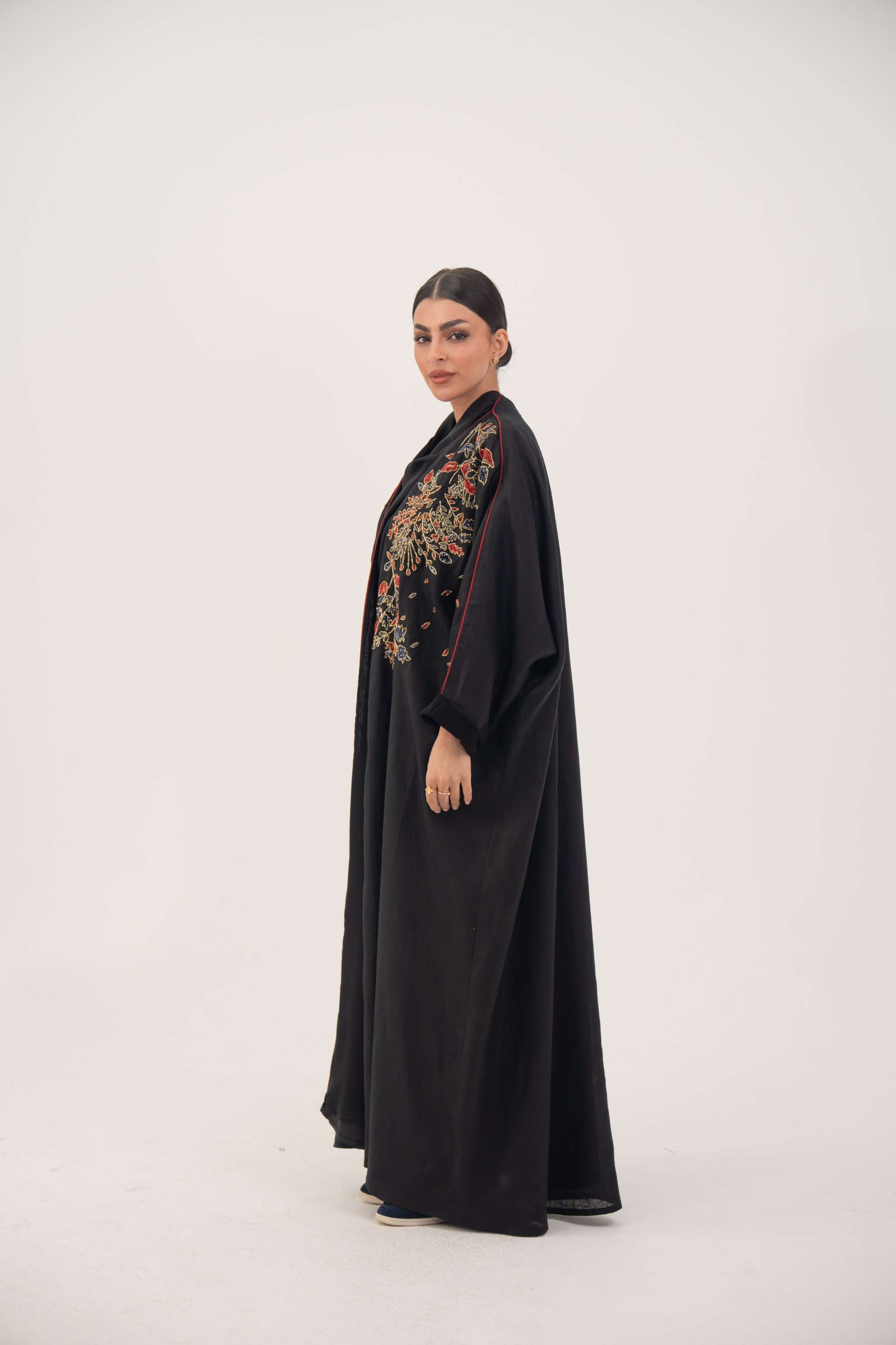 Woman wearing premium black linen abaya with multicolored red, gold, and navy shak embroidery and red trim, loose flowing bisht silhouette - abaya crafted from premium black linen