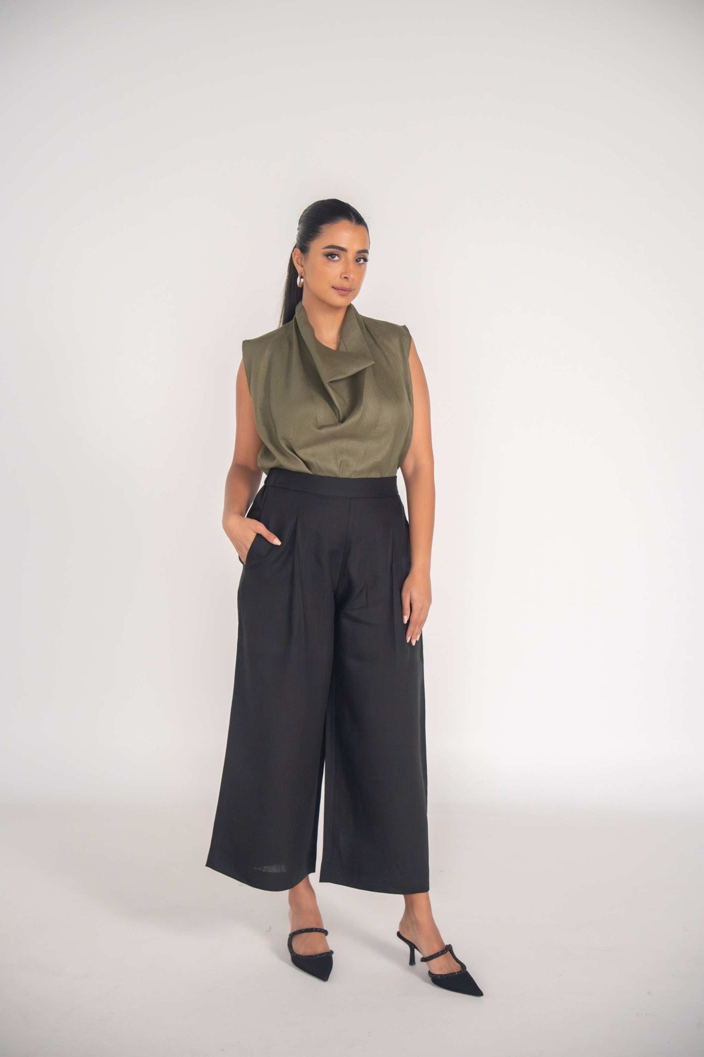 Woman wearing olive-green Raya Blouse sleeveless linen top paired with black wide-leg pants and black heels.