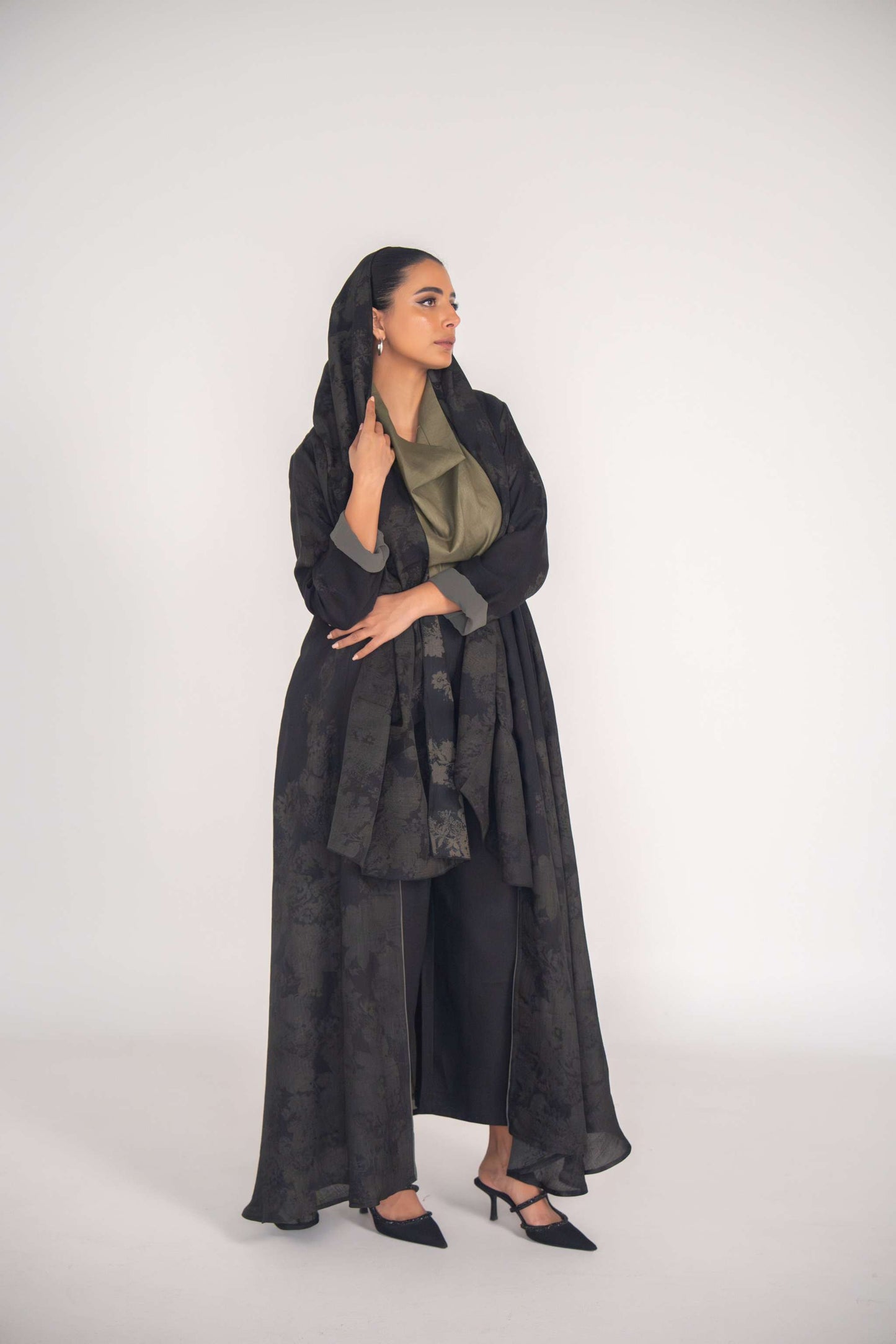 Woman wearing elegant black jacquard abaya with subtle floral pattern and grey cuff accents, styled with black pants and heels.