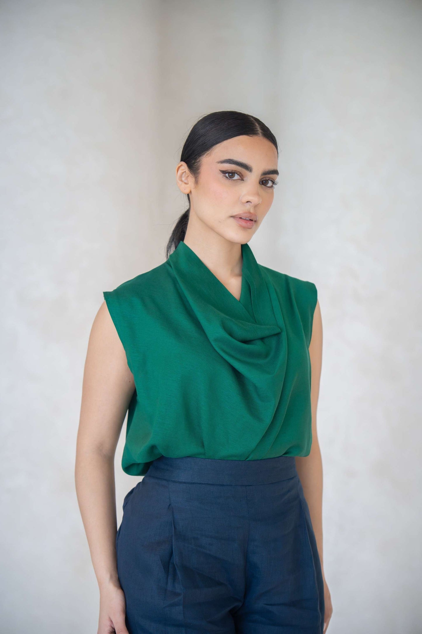 Woman wearing emerald green sleeveless blouse with cowl neckline and navy blue pants against neutral background
