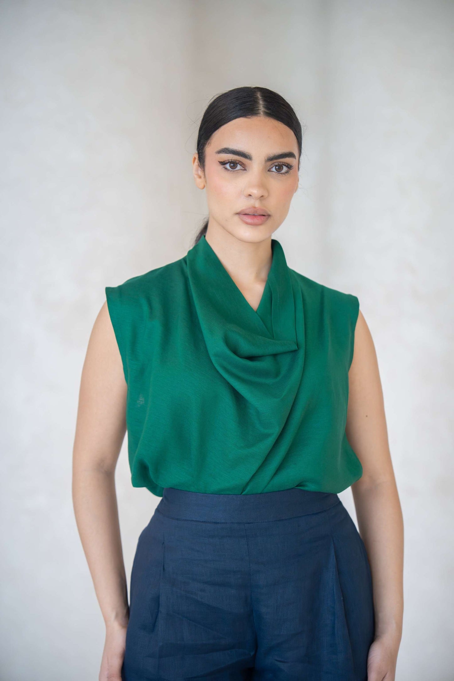 Woman wearing emerald green sleeveless blouse with cowl neckline and navy blue pants against a neutral background