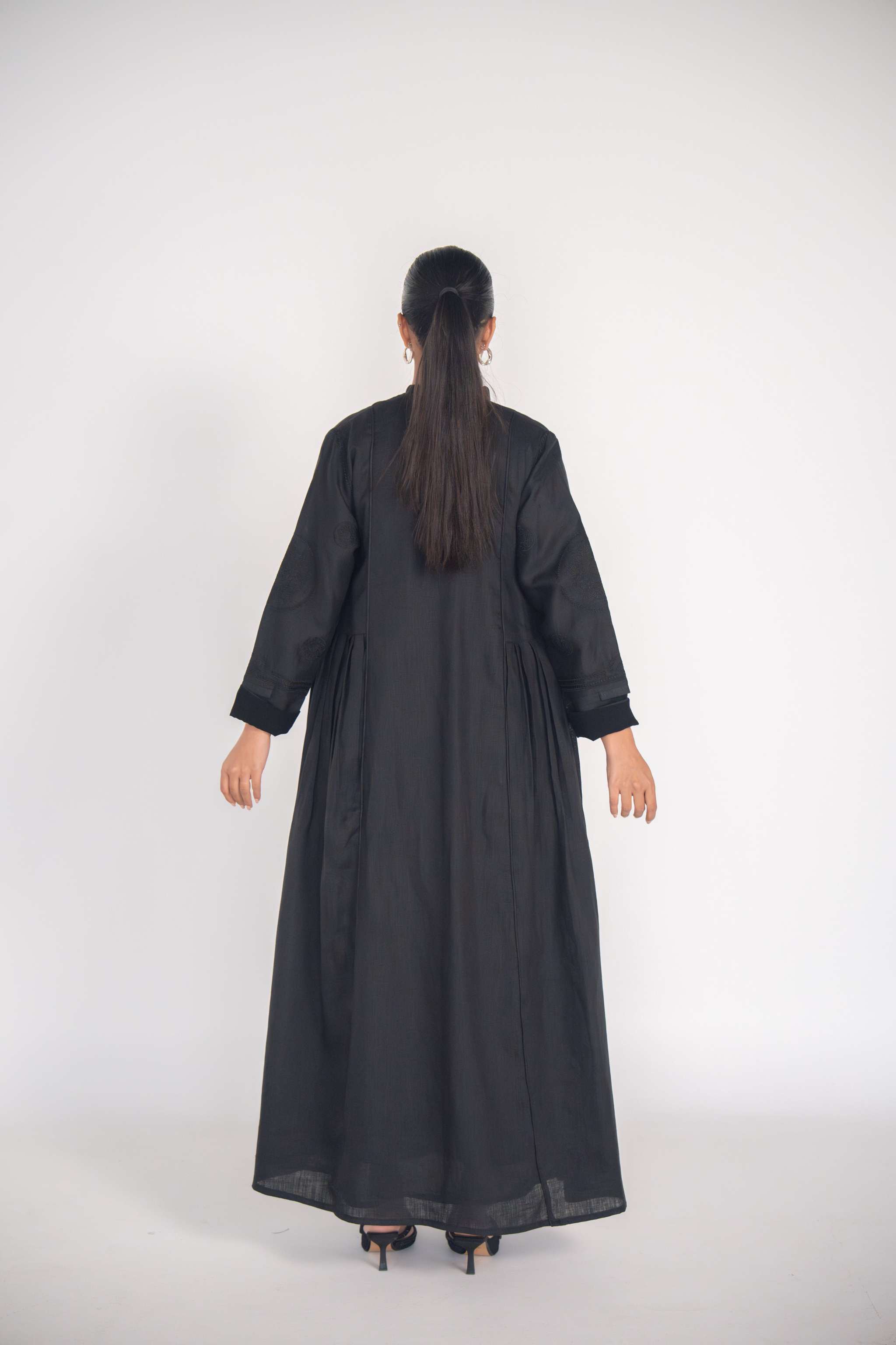 Back view of a woman wearing a refined black linen abaya with soft pleats and subtle black-on-black embroidery, featuring full lining and a flowy silhouette.