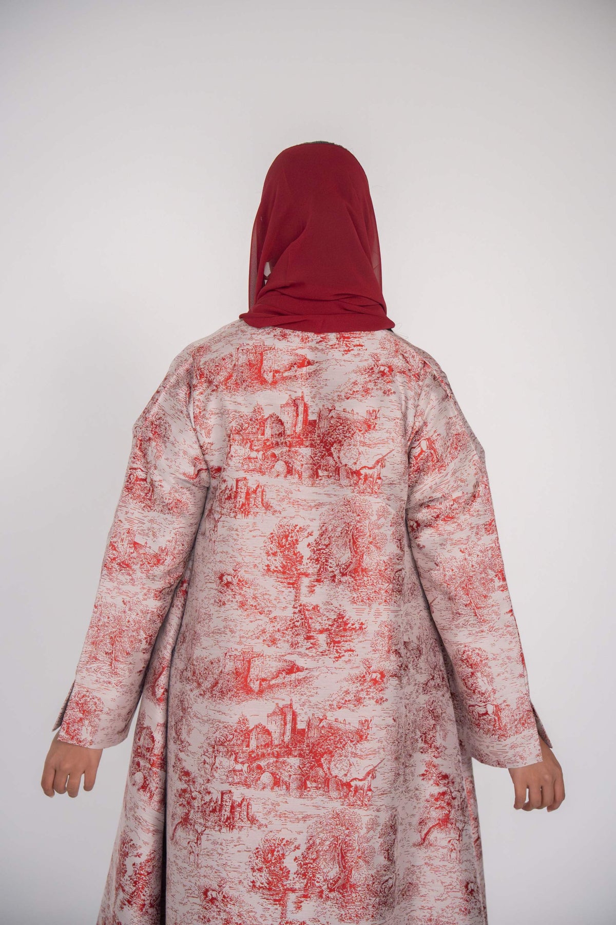 Back view of luxurious jacquard abaya with red Toile de Jouy countryside pattern on off-white fabric, worn with red headscarf.