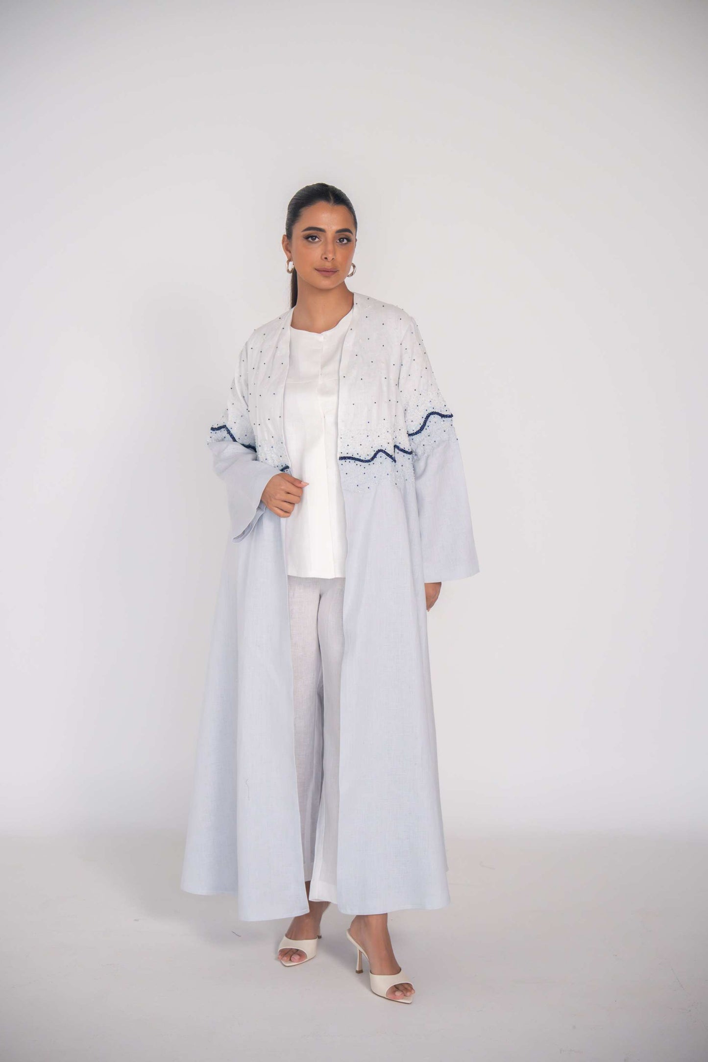 Woman wearing a light linen abaya with hand-applied blue and white beadwork in a wave pattern on bodice and sleeves, featuring a flowing cut and chiffon lining.