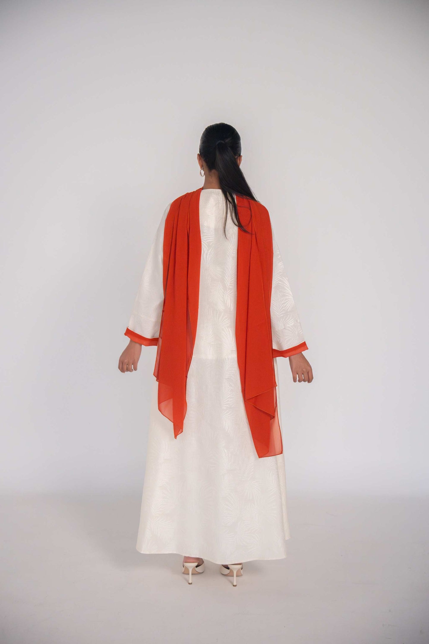 Woman wearing a white abaya made of textured jacquard fabric with an orange chiffon scarf draped over her shoulders, shown from the back.
