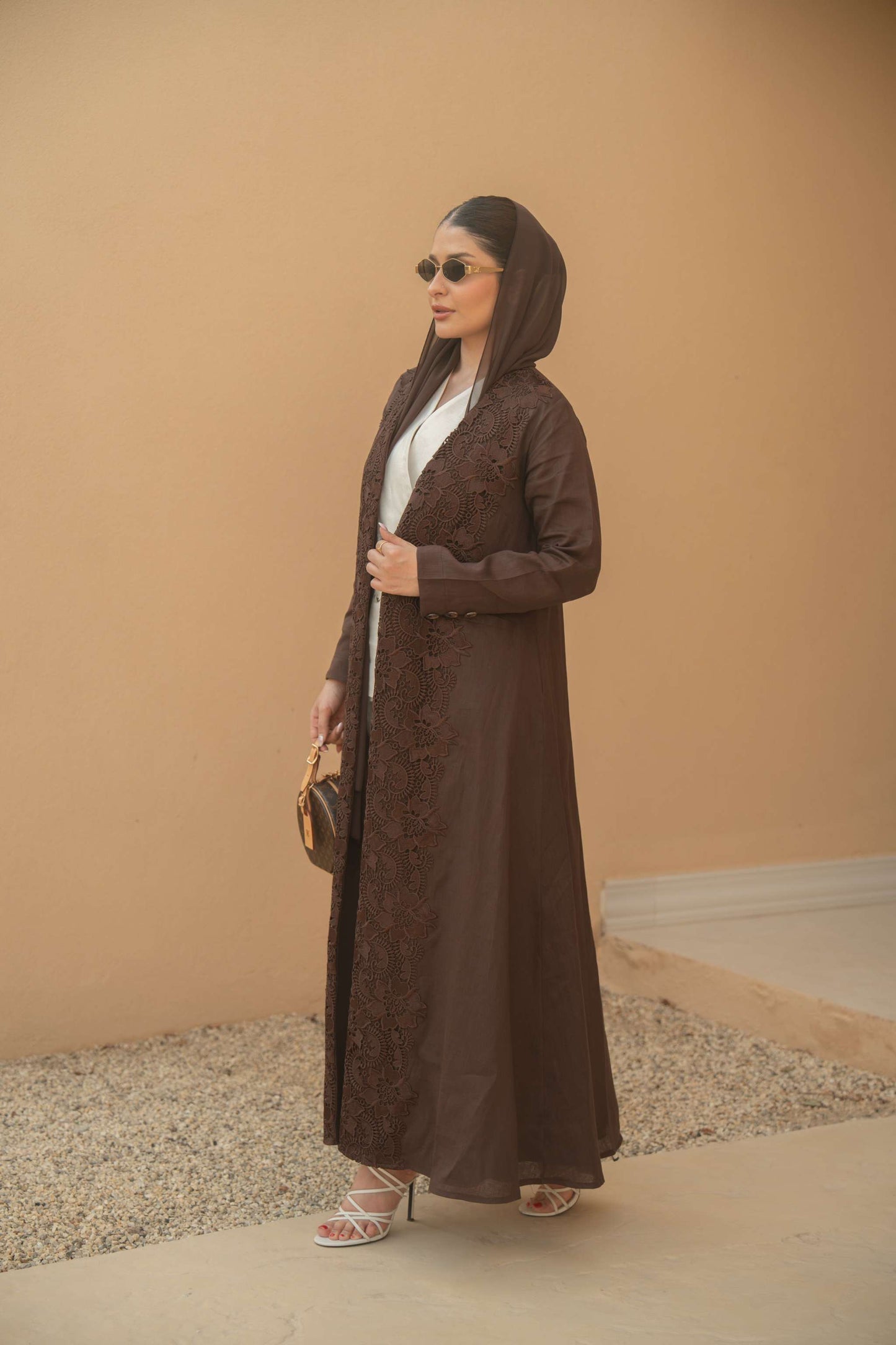 Woman wearing deep mocha brown Mocha Bloom Abaya with floral embroidery and open front design, styled with sunglasses and heels against neutral background