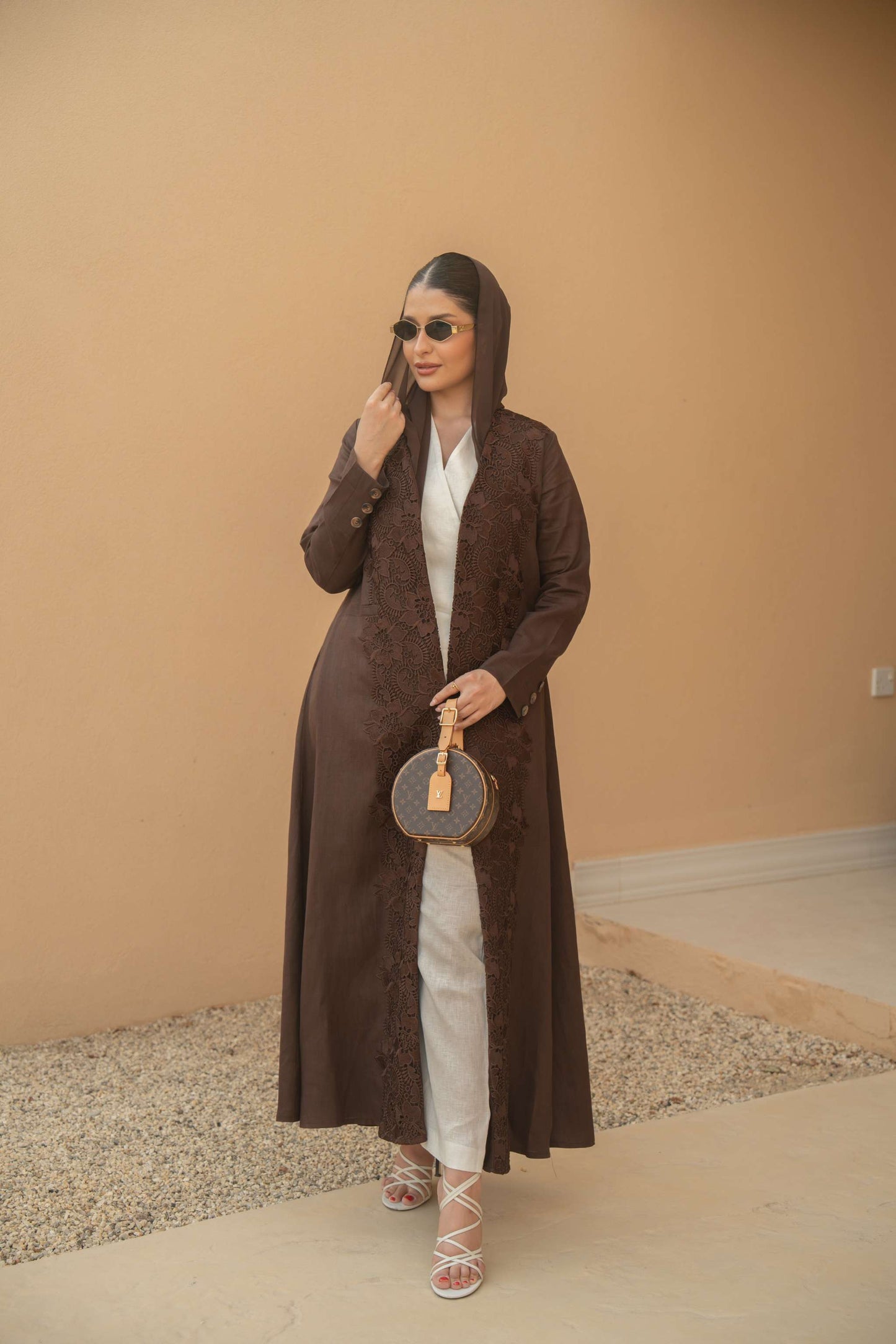 Woman wearing a deep mocha brown Mocha Bloom Abaya with floral embroidery, paired with white outfit and sunglasses, holding a round handbag.