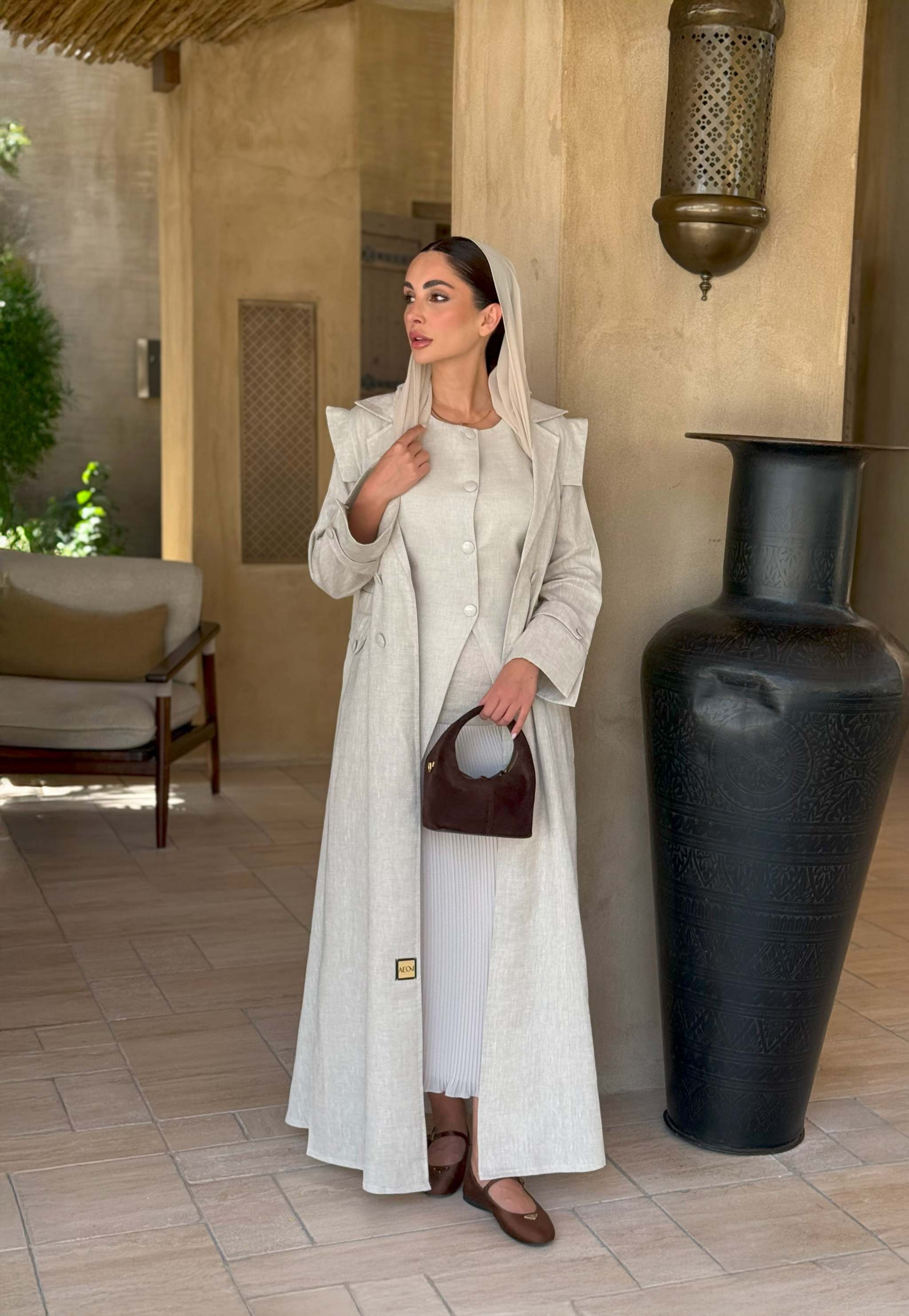 woman wearing a beige linen abaya with trench-style silhouette and matching sheila holding a brown handbag - beige abaya