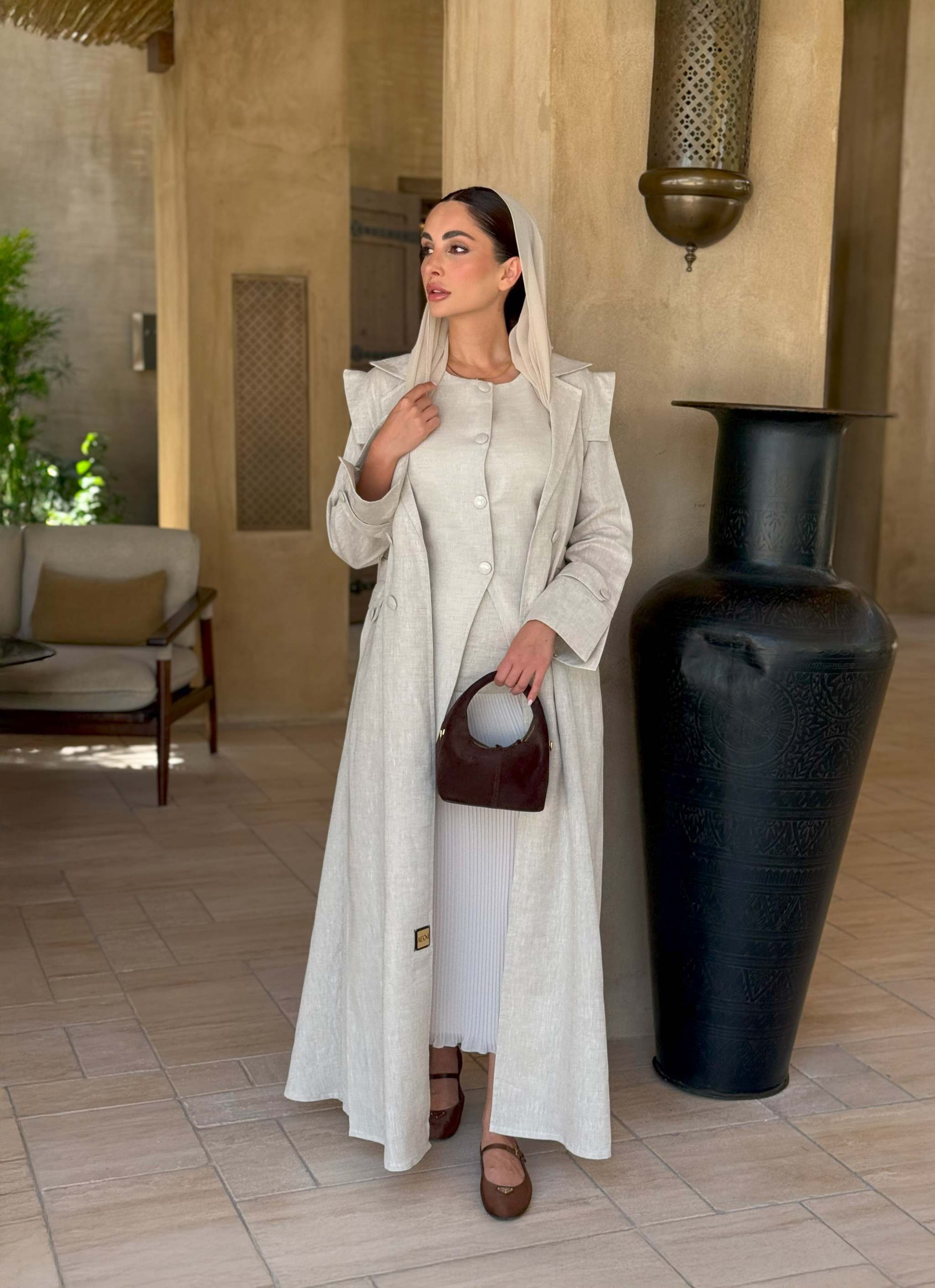 woman wearing a beige linen abaya with trench-style silhouette and matching sheila holding a brown handbag - beige abaya