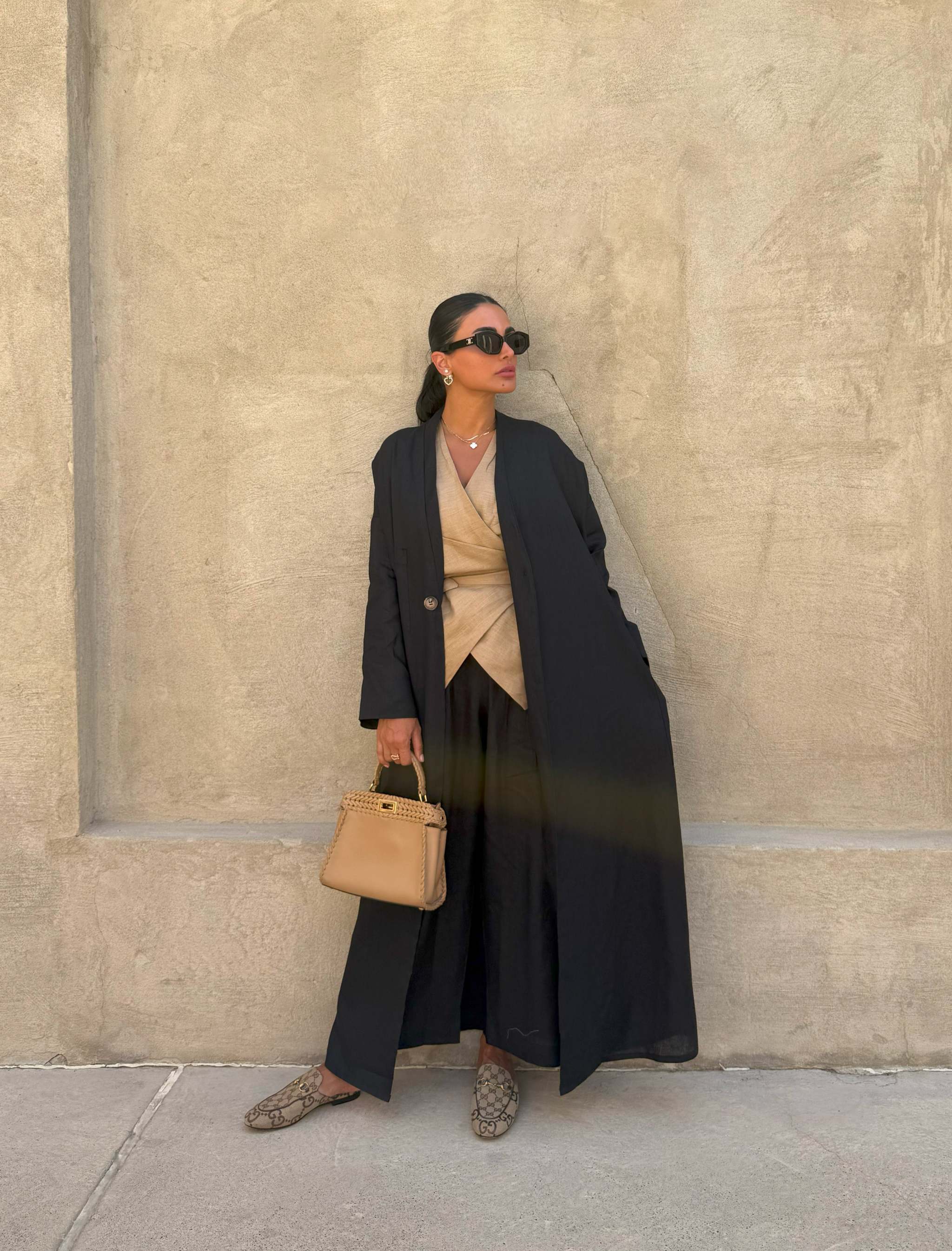 Woman wearing a flowy black Midnight Linen abaya over beige wrap top and wide-leg pants, accessorized with sunglasses and a tan handbag.