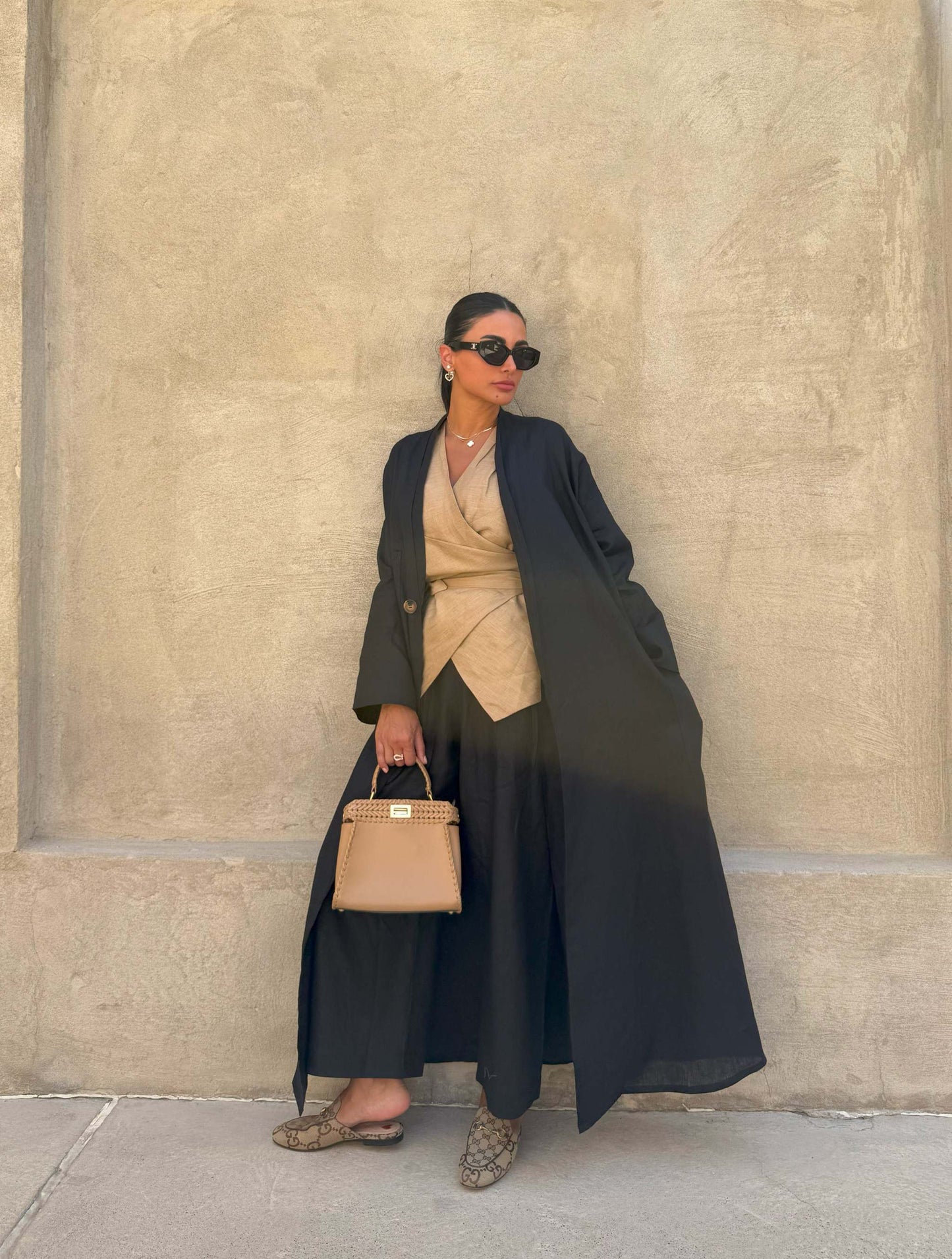 Woman wearing minimalist black linen abaya with straight cut and open front, paired with beige outfit and sunglasses against beige wall