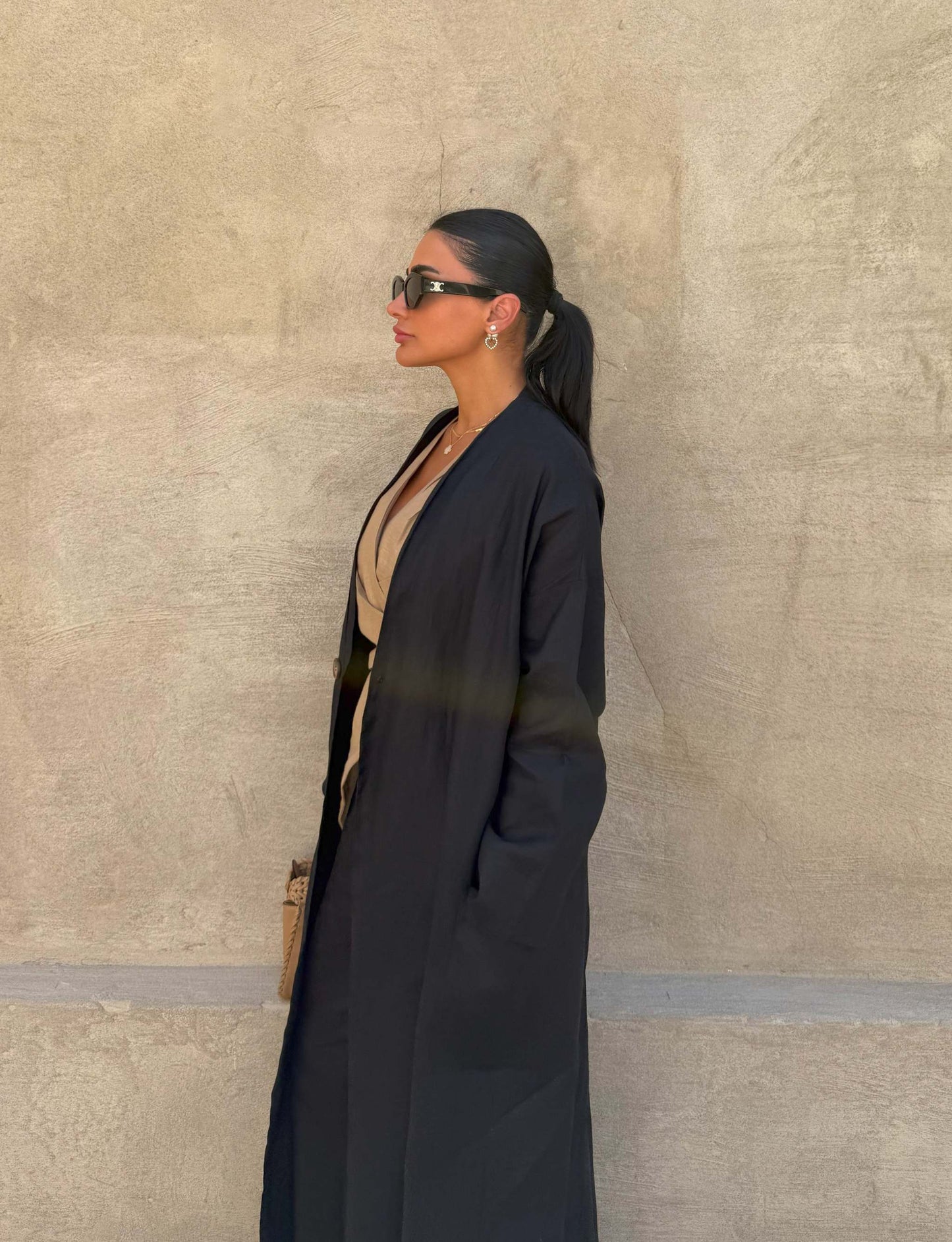 Woman wearing minimalist black linen abaya with straight cut and open front against beige wall