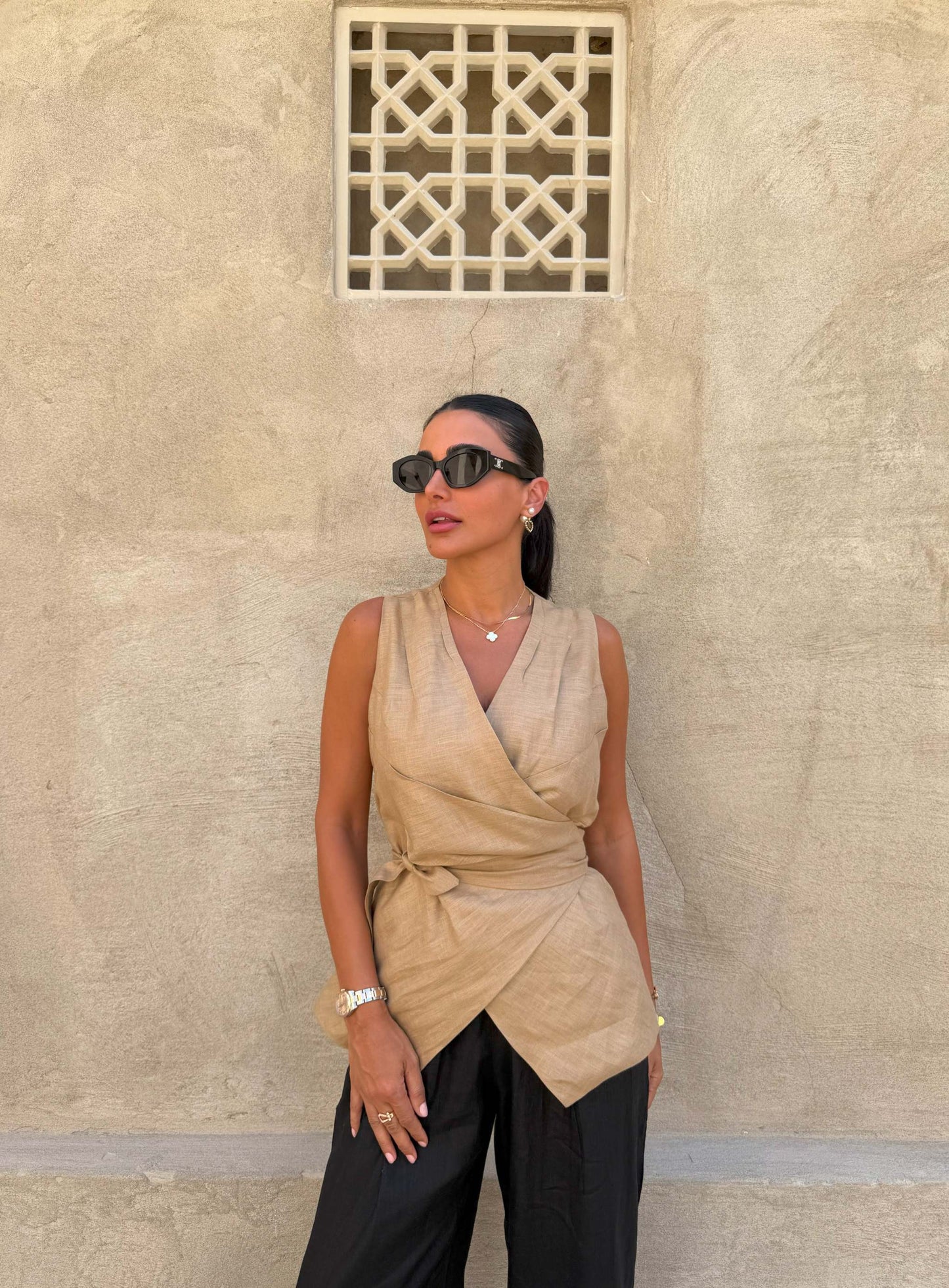 Woman wearing a sleeveless beige Clay Vest wrap top with a waist tie and black pants against a textured beige wall background