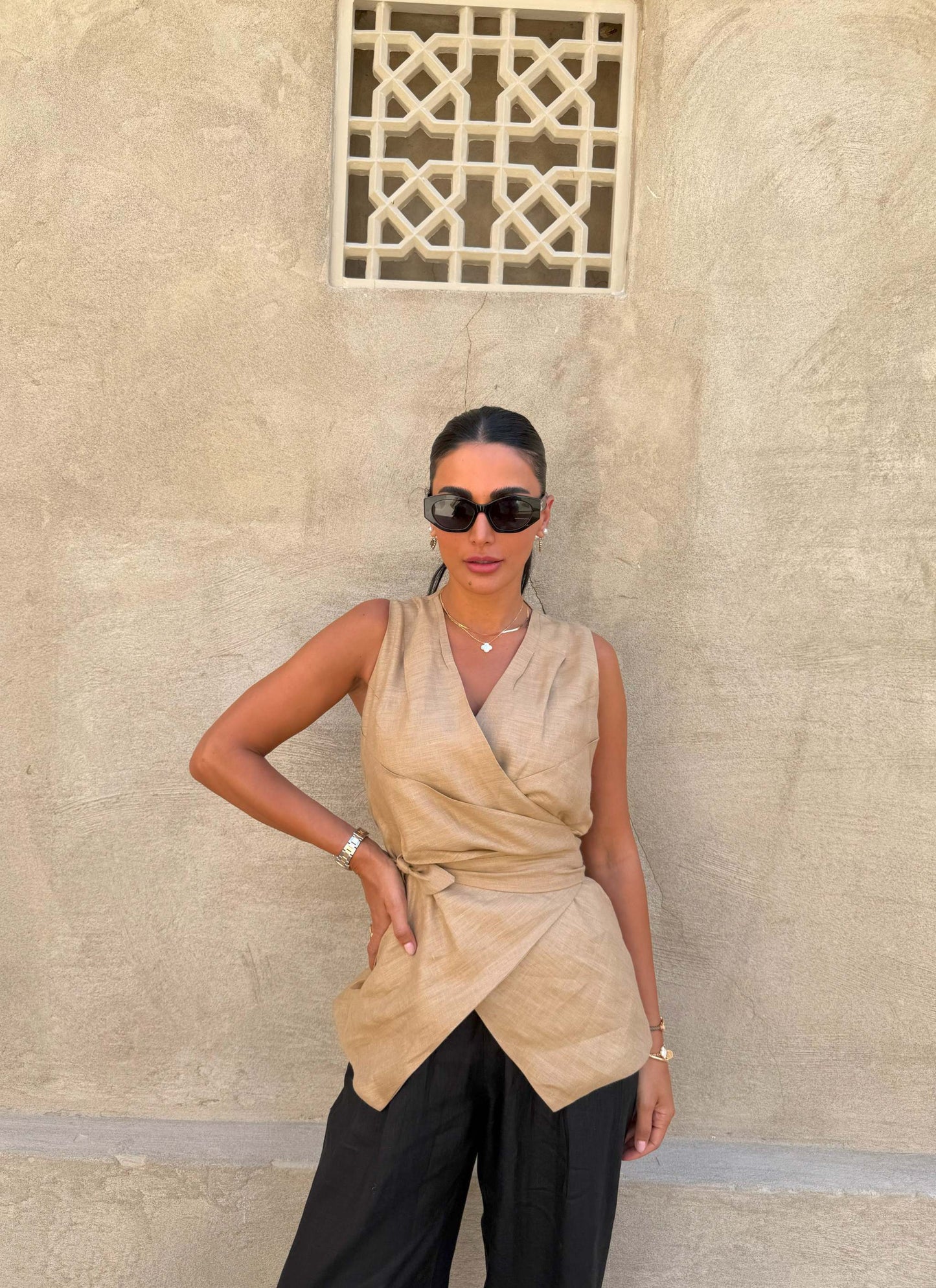 Woman wearing sleeveless beige clay vest with waist tie and black pants against textured wall background