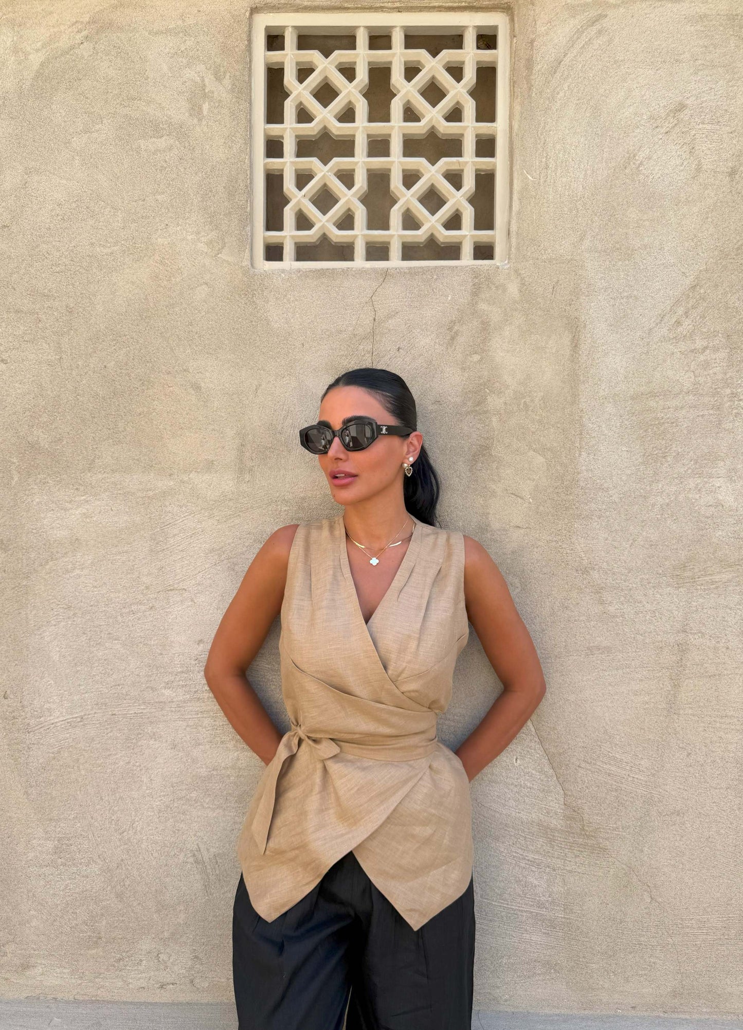 Woman wearing sleeveless beige clay vest with waist tie and black pants against textured wall background