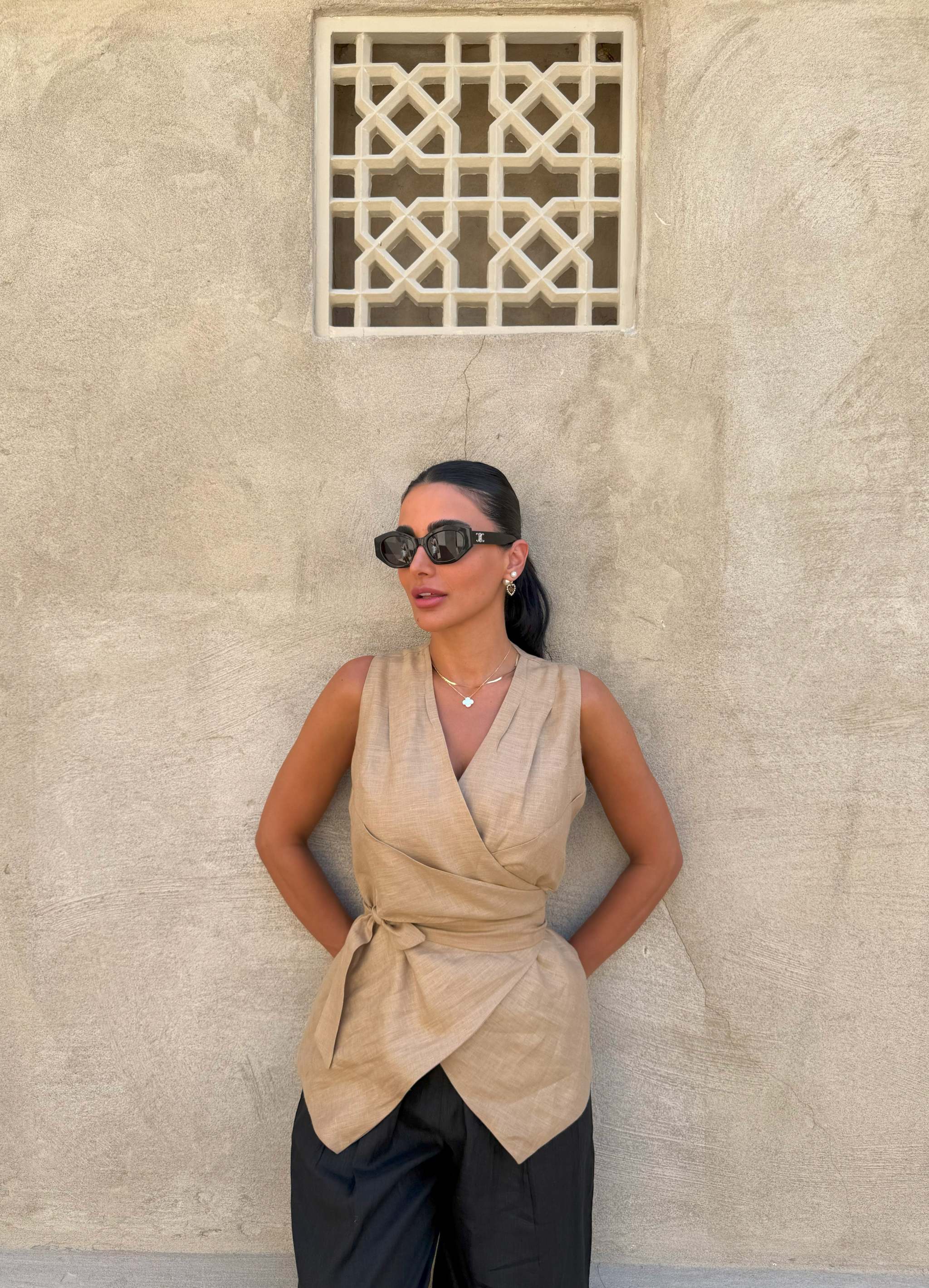 Woman wearing sleeveless beige clay vest with waist tie and black pants against textured wall background