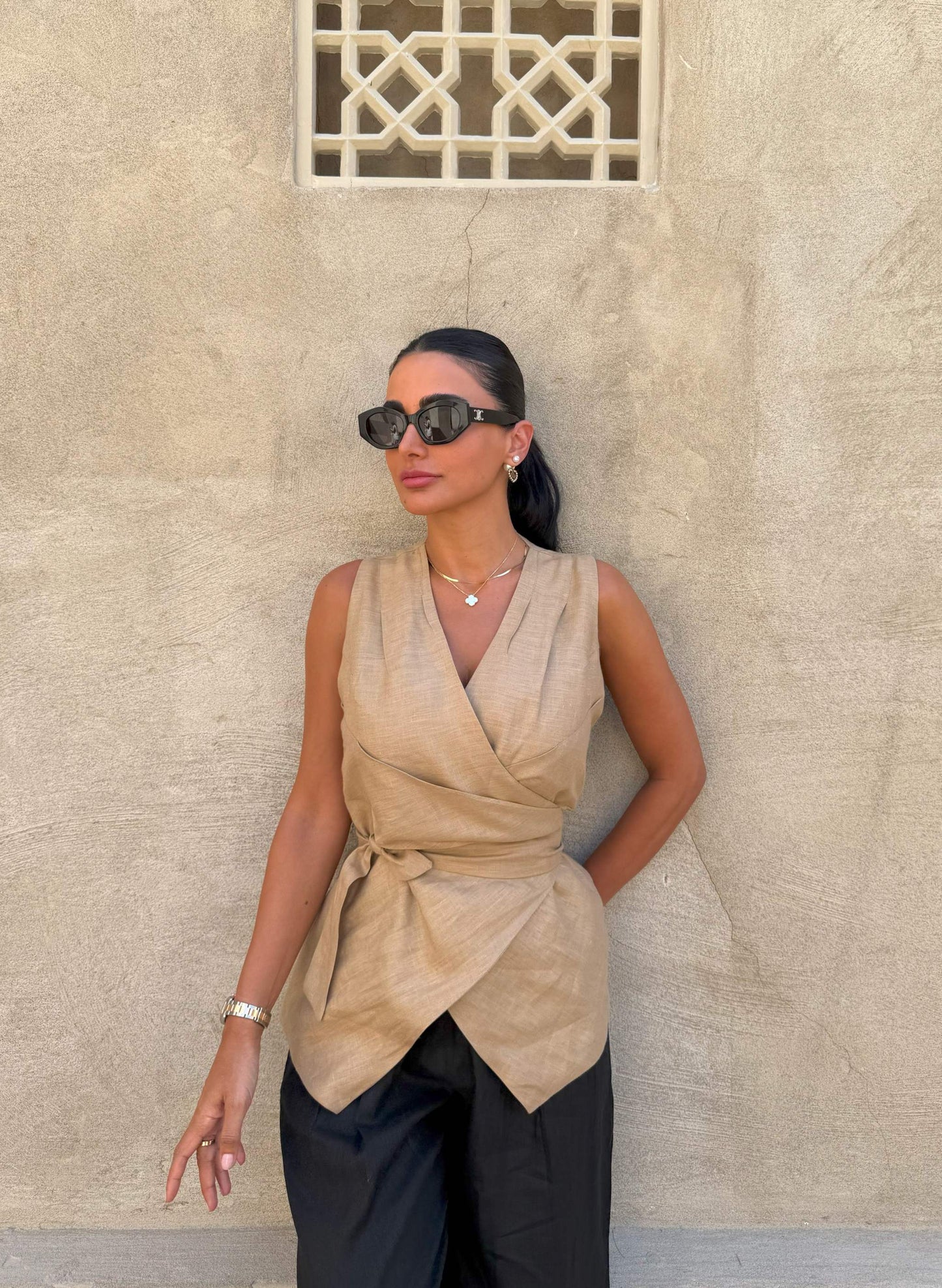 Woman wearing sleeveless beige clay vest with waist tie and black pants against textured wall background