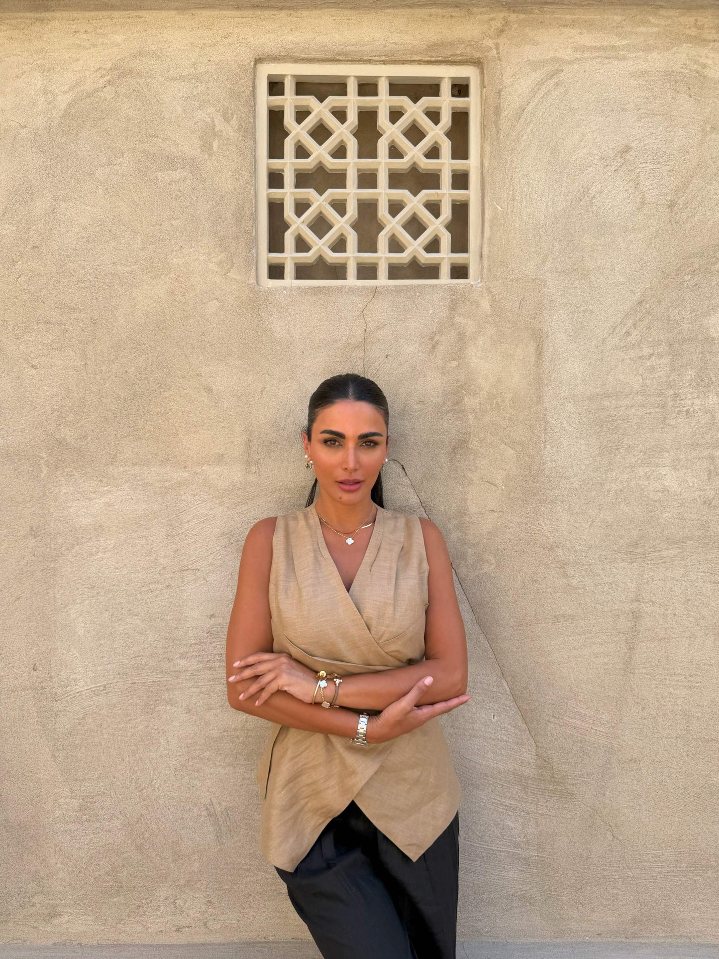 Woman wearing sleeveless beige clay vest with waist tie and black pants against textured wall background