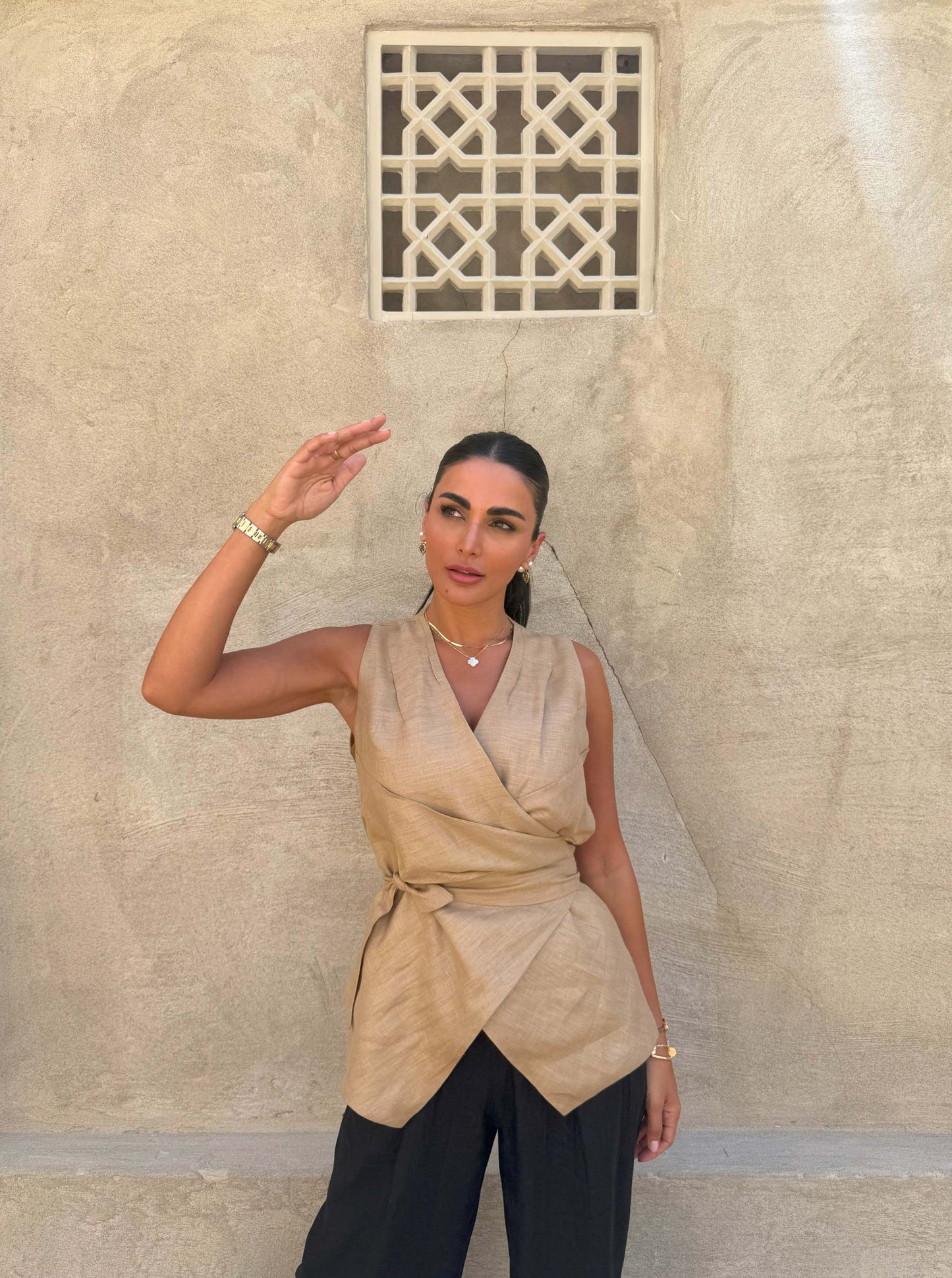Woman wearing sleeveless beige clay vest with waist tie and black pants against textured wall background