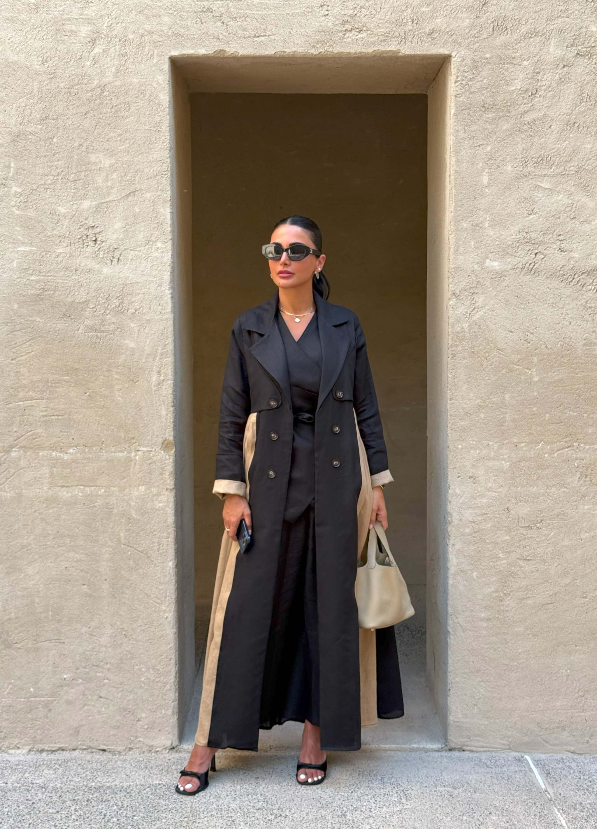 Woman wearing Latte Smoke trench-style black abaya with beige side panels and cuffs, double-breasted design and structured collar, holding beige handbag.