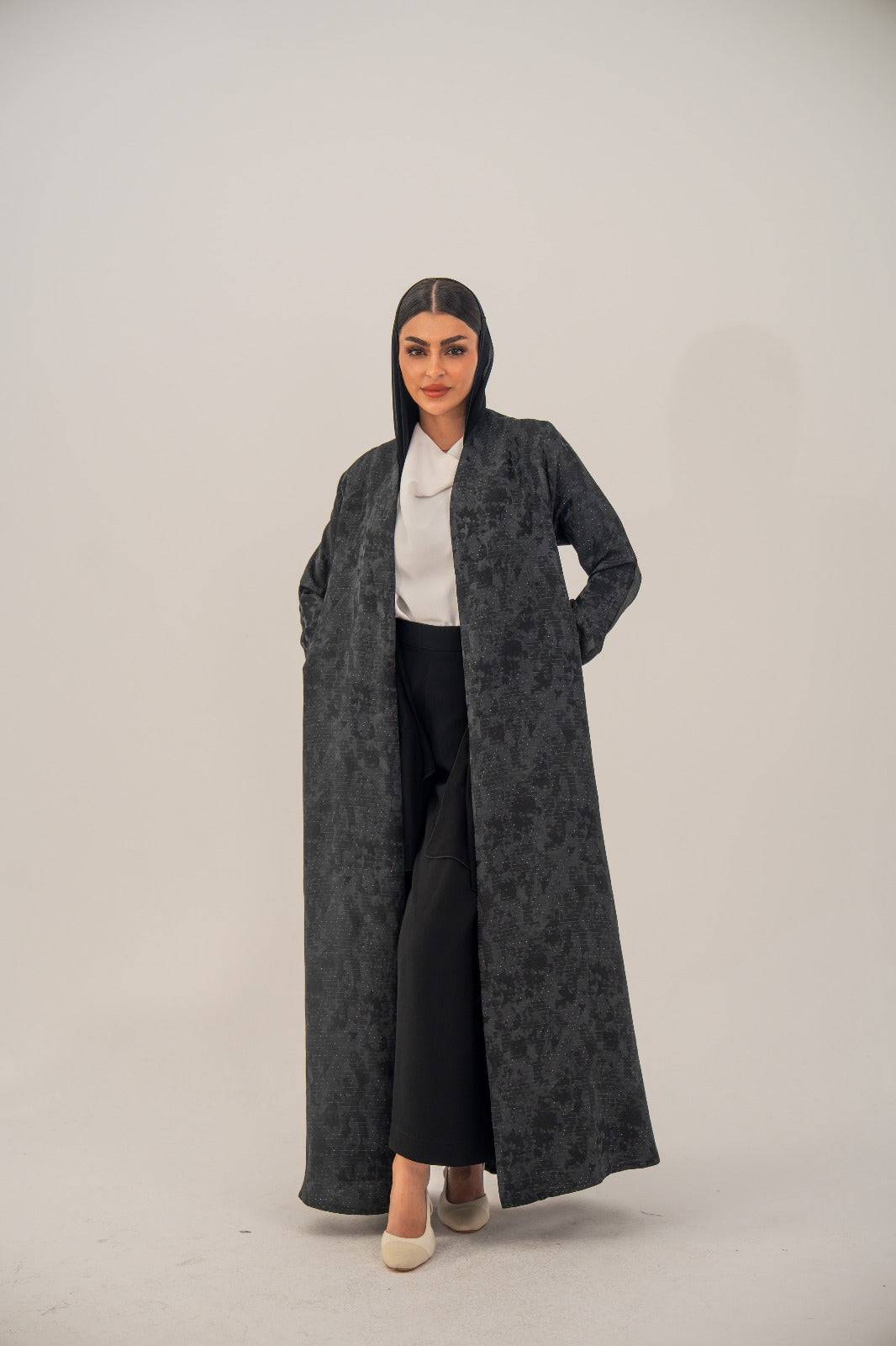 Woman wearing an open-front black silk abaya with subtle abstract pattern and light shimmer, styled with white top and black pants.