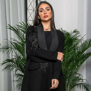 Woman wearing a black crepe blazer-style abaya with satin lapel and feather detailing on one shoulder.