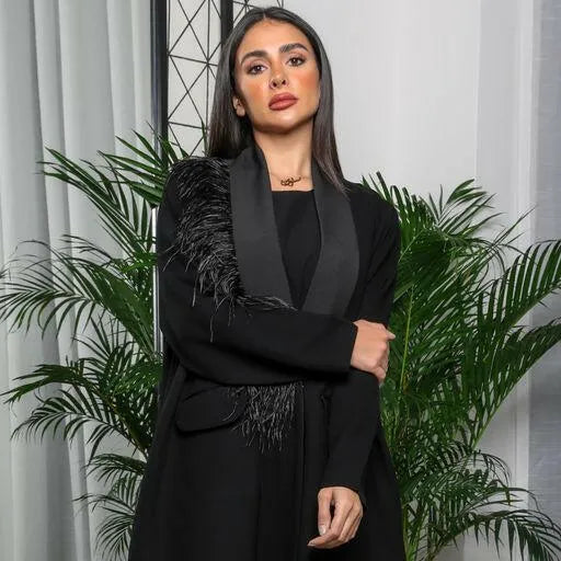 Woman wearing a black crepe blazer-style abaya with satin lapel and feather detailing on one shoulder.