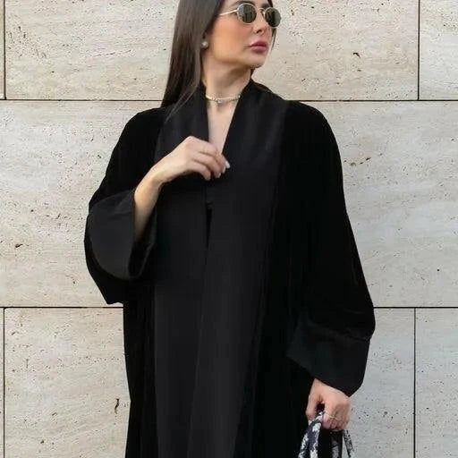 Woman wearing Crepe Dusk black velvet bisht with crepe accents and freesize elegant flowing cut against stone wall background