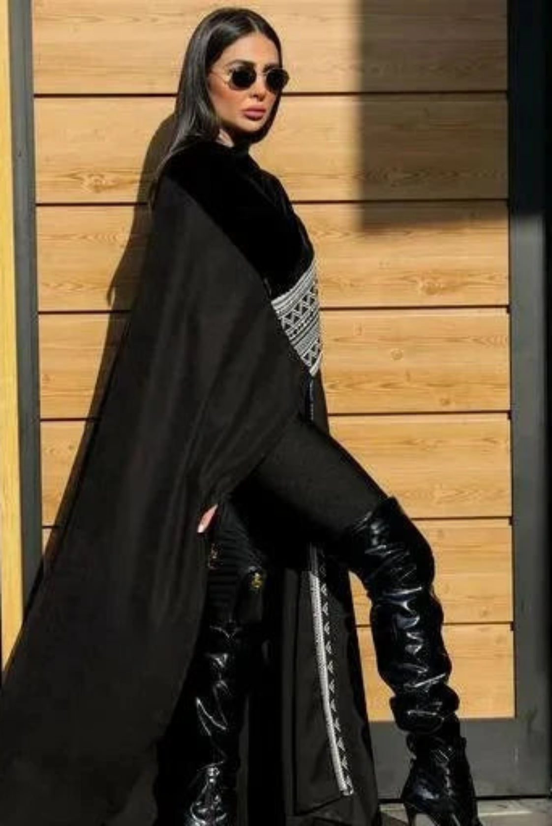 Woman wearing Midnight Veil black chamois and velvet cape with thigh-high boots and sunglasses standing against wooden wall