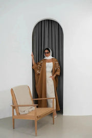 Woman wearing camel brown bisht-style abaya with hand embroidery standing by an arched doorway next to a wooden chair