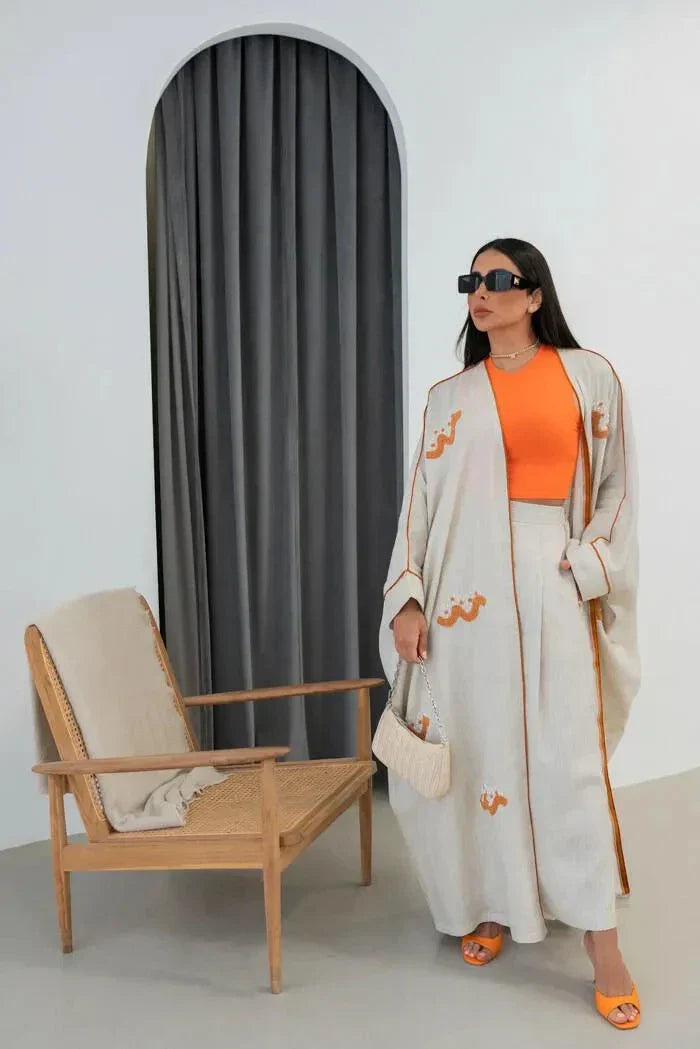 Woman wearing beige linen abaya with orange hand-embroidered Arabic script motifs and orange inner top standing beside wooden chair