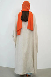 Woman wearing beige linen abaya with orange hand-embroidered Arabic script motifs and orange inner top standing beside wooden chair