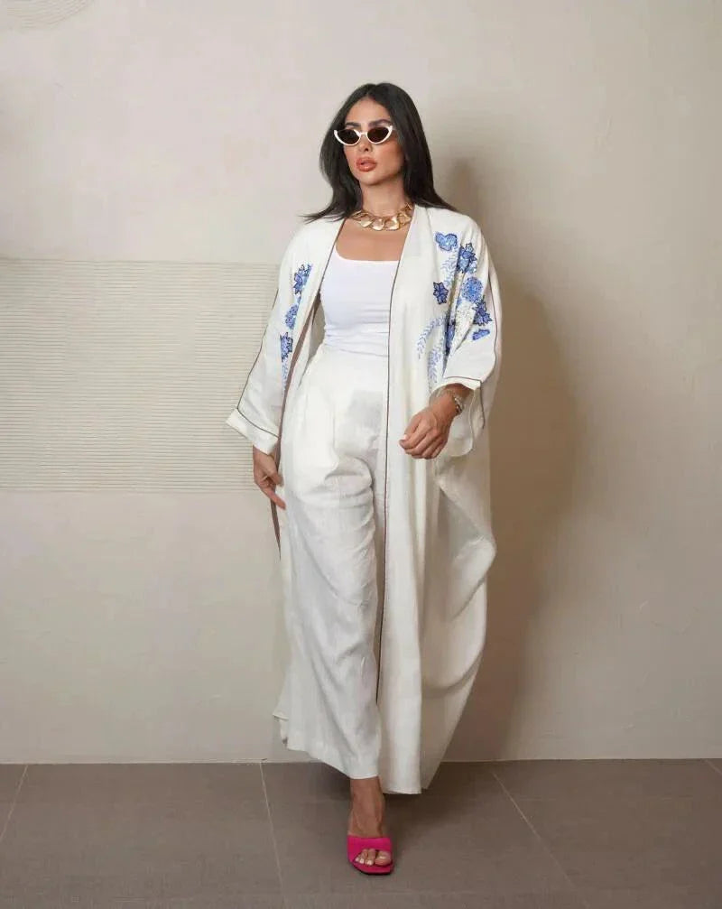 Elegant white abaya from linen with blue and gold hand embroidery, loose flowing silhouette, styled with white pants and pink heels
