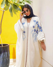 Elegant white abaya from linen with blue and gold hand embroidery, loose flowing silhouette, styled with white pants and pink heels