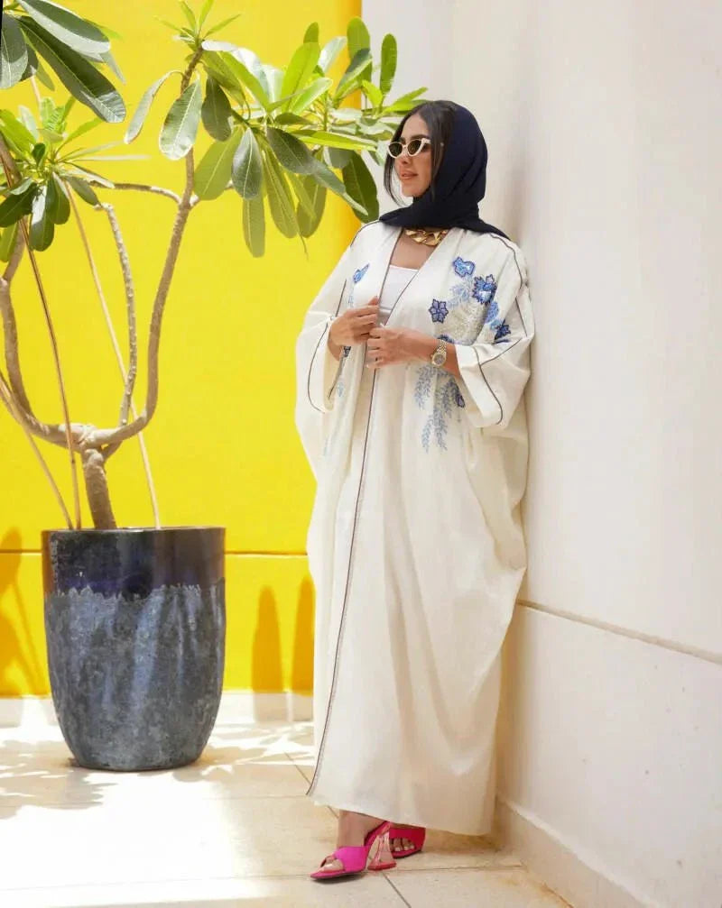 Elegant white abaya from linen with blue and gold hand embroidery, loose flowing silhouette, styled with white pants and pink heels