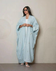 Woman wearing a sky-blue lightweight linen abaya with delicate white and crystal hand embroidery, styled with white sandals and sunglasses.