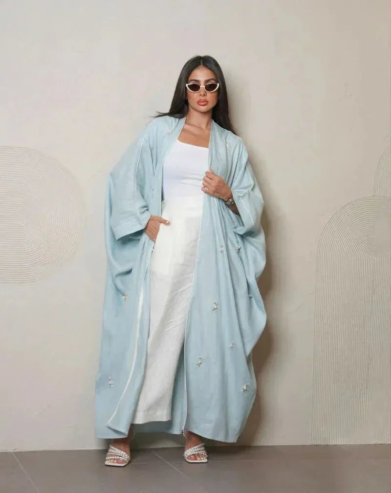 Woman wearing a sky-blue lightweight linen abaya with delicate white and crystal hand embroidery, styled with white sandals and sunglasses.